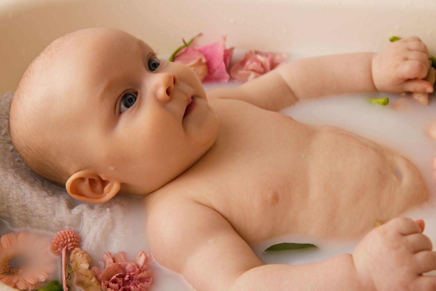 A baby lies on white bedding surrounded by delicate pink rose petals creating a sweet and dreamy portrait.
