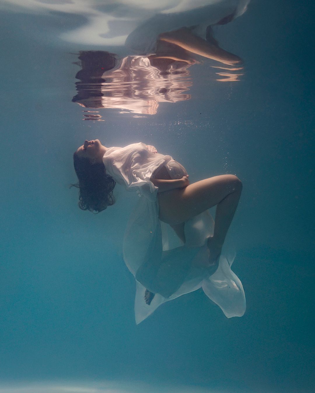 Pregnant woman in white flowing dress floating gracefully underwater in a serene blue pool, maternity photography.