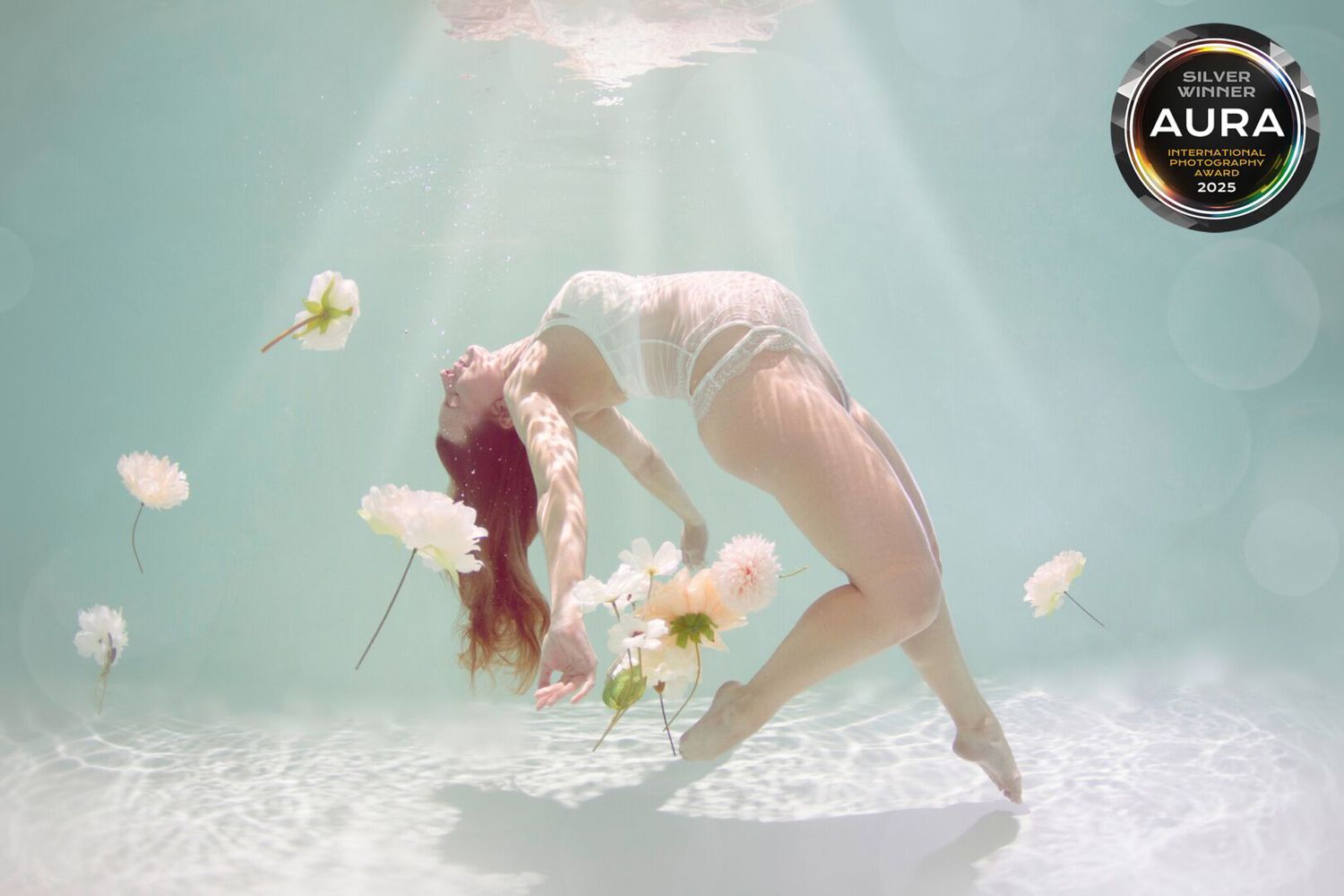 A person in a white dress floats gracefully underwater surrounded by delicate white flowers and bubbles.