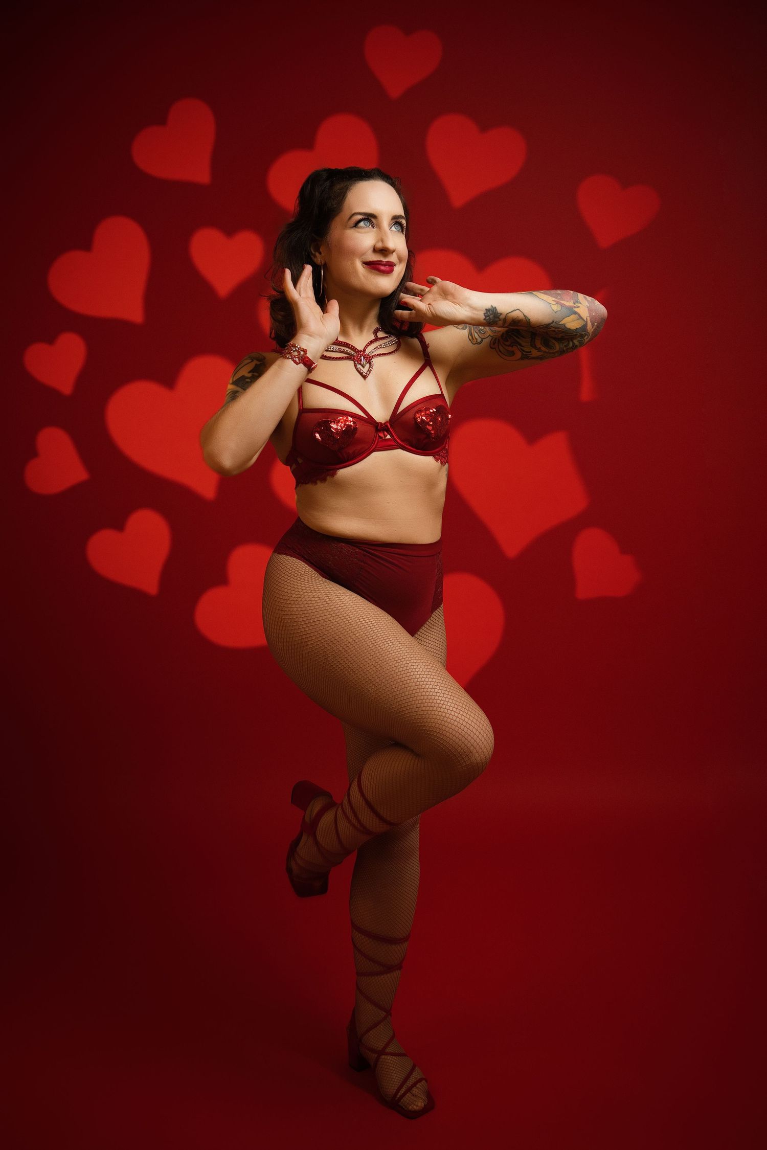 Woman in red sequin lingerie poses against red background with heart shapes, showcasing tattoos and strappy heels.