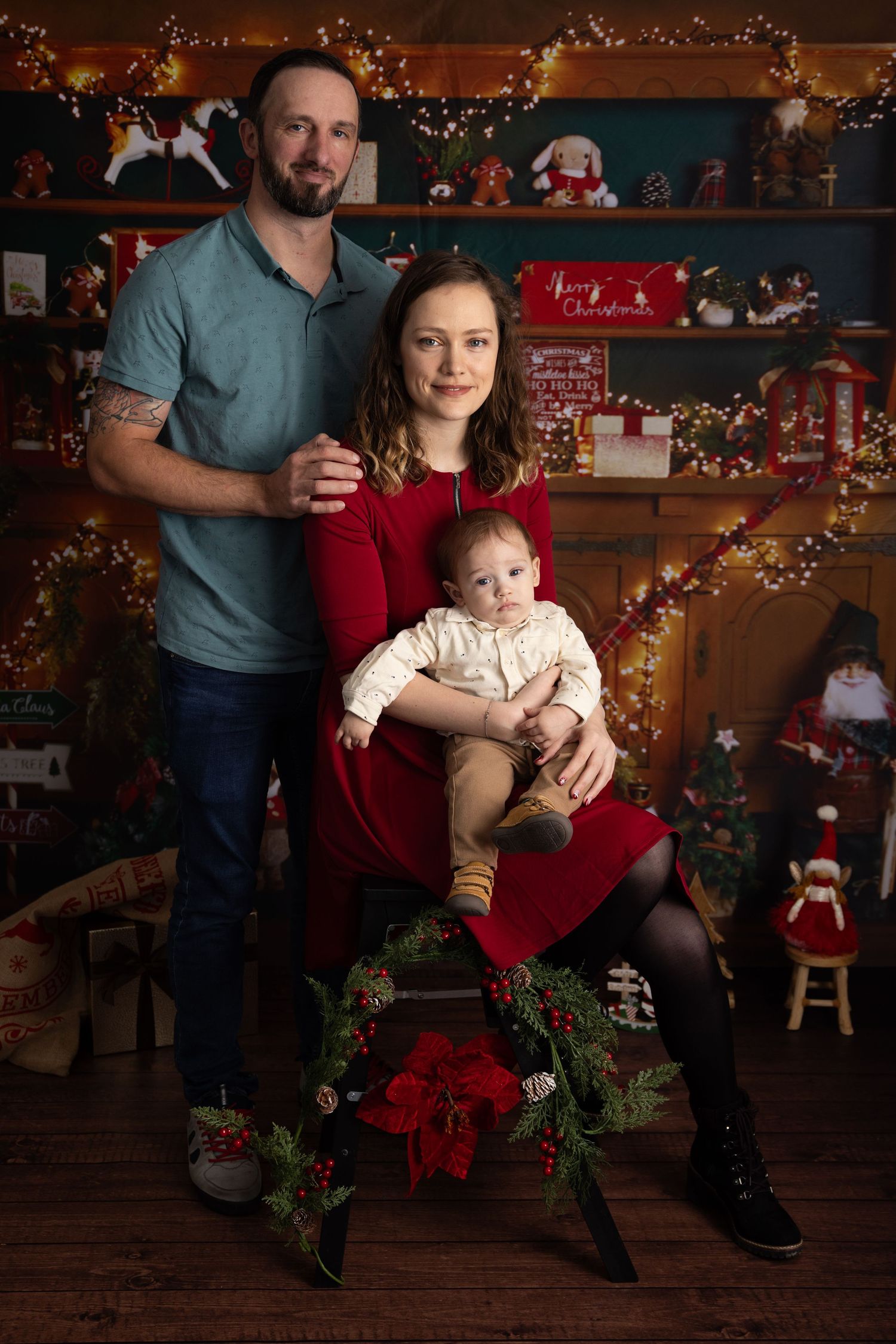 A family poses together against a rustic backdrop with twinkling Christmas lights and holiday decorations.