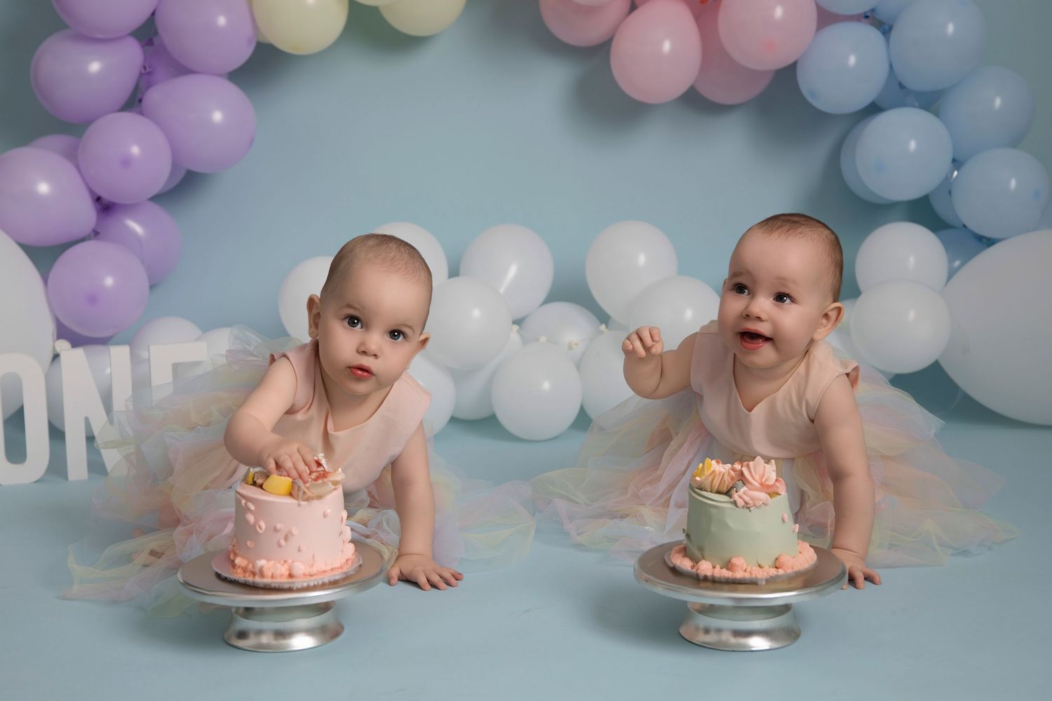 Two smiling infants at a cake smash photo session with pastel balloon backdrop and small individual cakes.