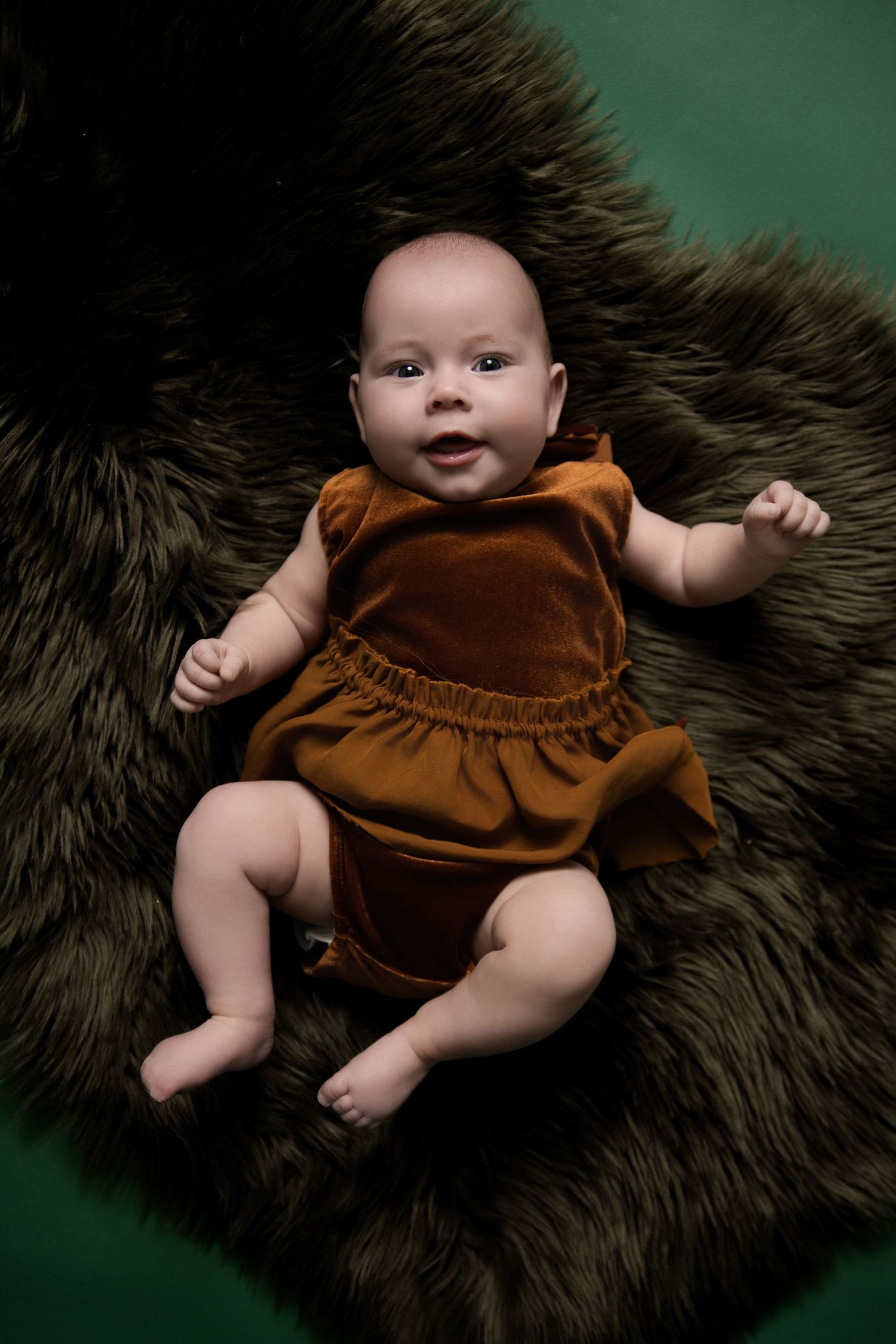 A baby wearing a brown velvet romper lies on a dark furry rug against a green background.