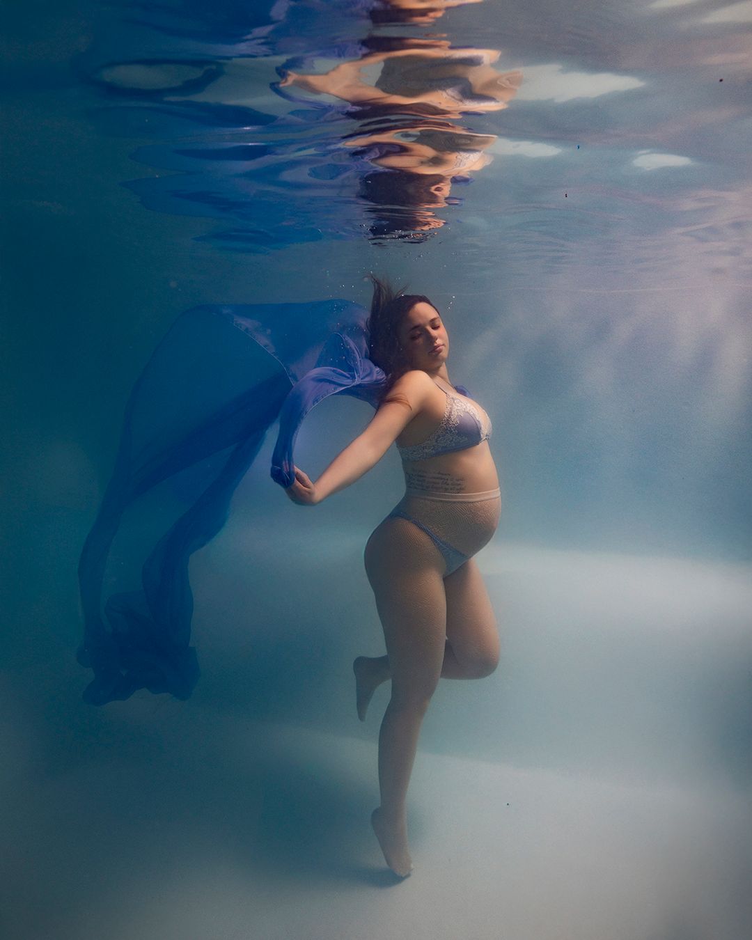 Pregnant woman underwater in blue bikini holding flowing blue fabric in a serene maternity photoshoot.