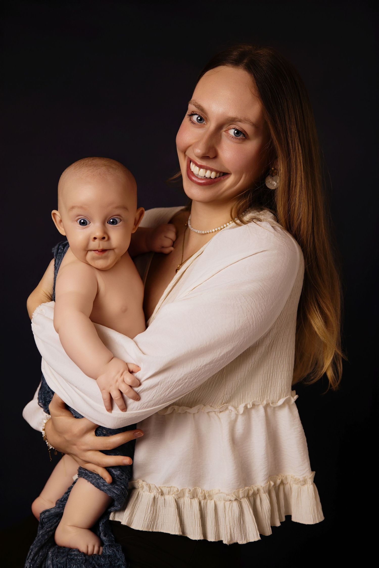 Mother in cream ruffled top holds smiling baby wearing navy shorts against dark background in studio portraits.