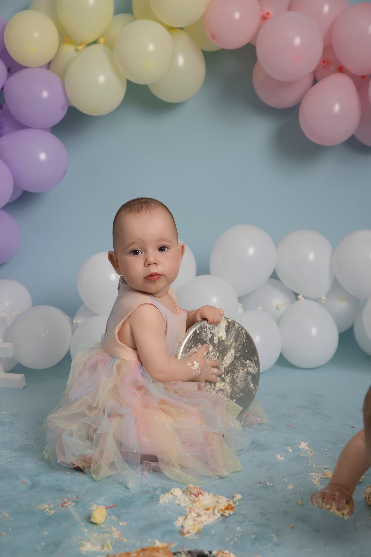 A baby in a tulle dress sits on a blue floor surrounded by pastel balloons and scattered cake crumbs for a cake smash photo.