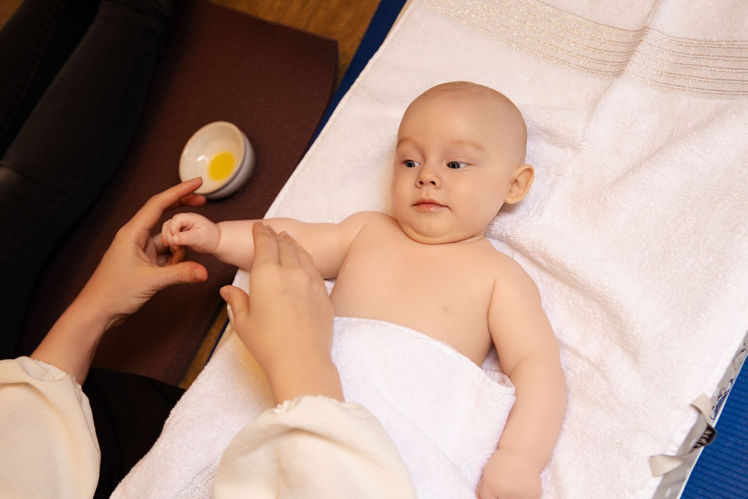 A baby lying on white sheets receives a vaccination shot from a medical professional's hand.