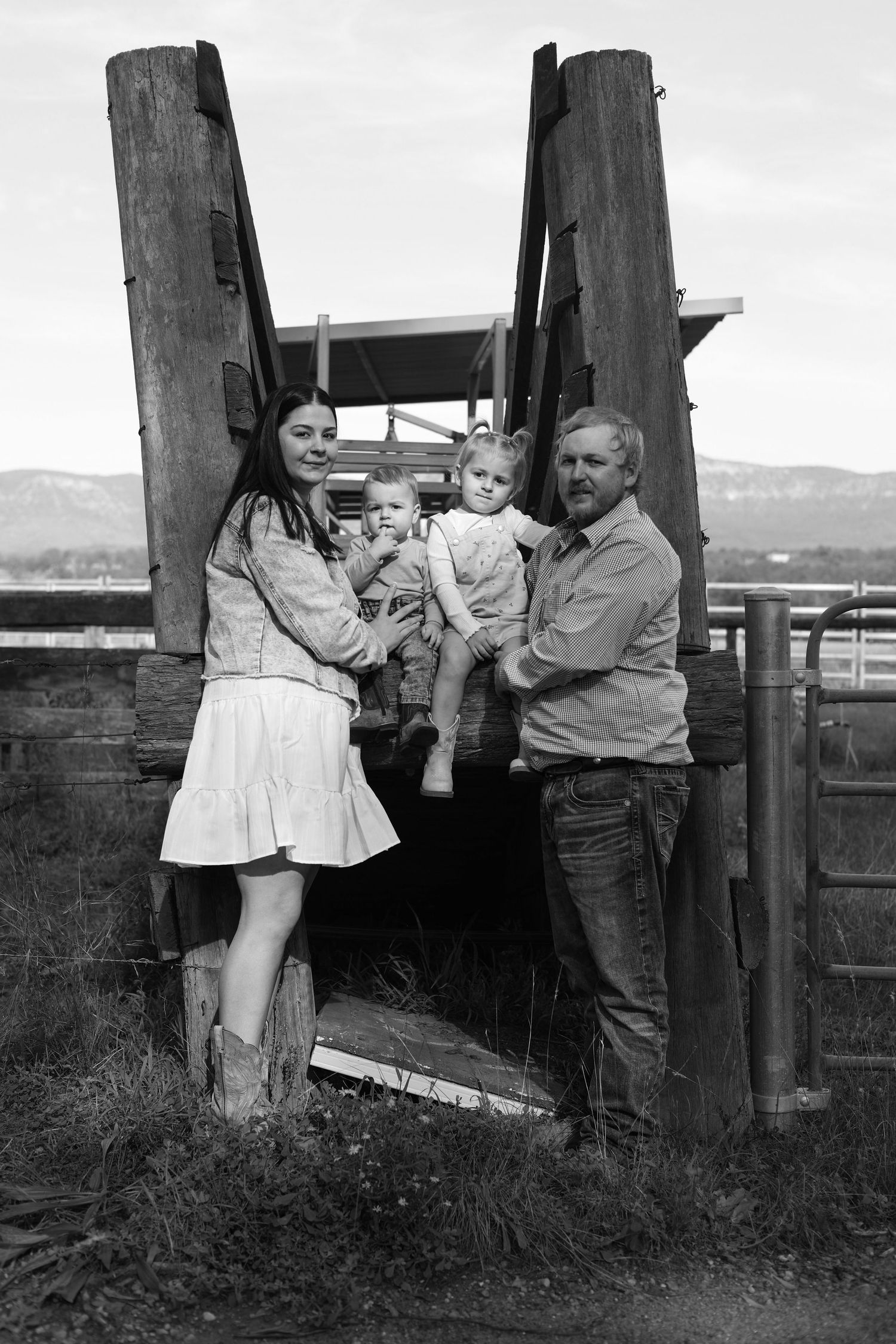 Black and white family portrait of parents with two toddlers at a rustic ranch gate, mountains in background.