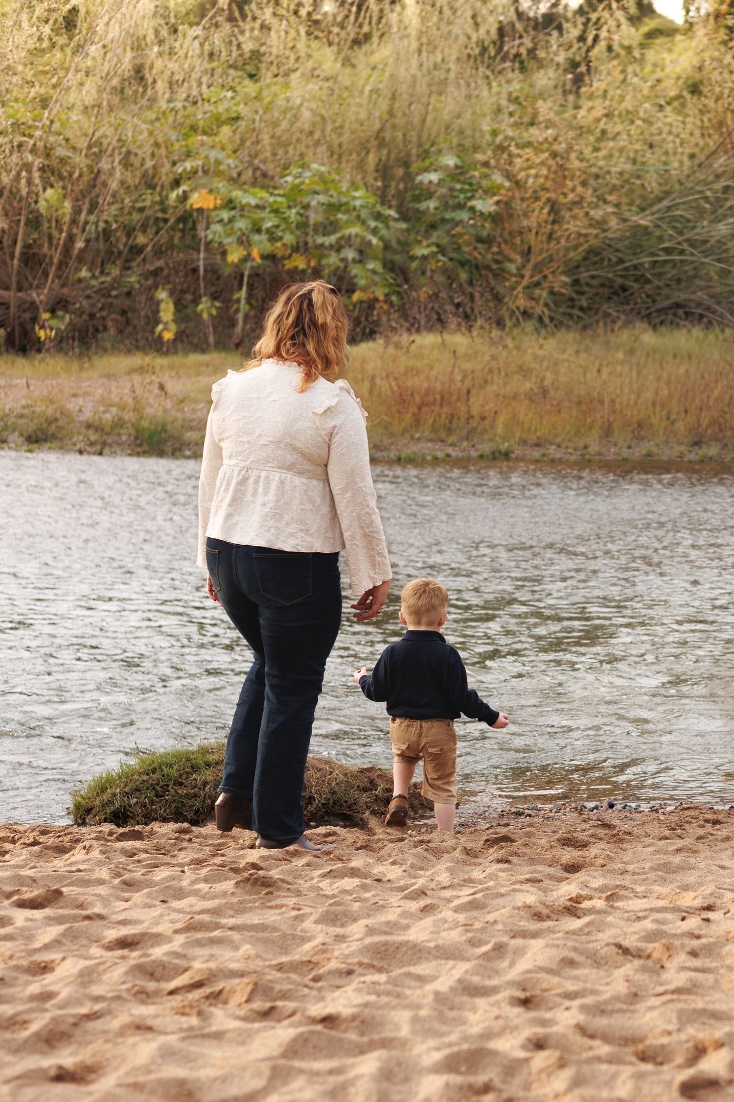 A mother and toddler walk hand-in-hand along a sandy riverbank surrounded by autumn foliage.
