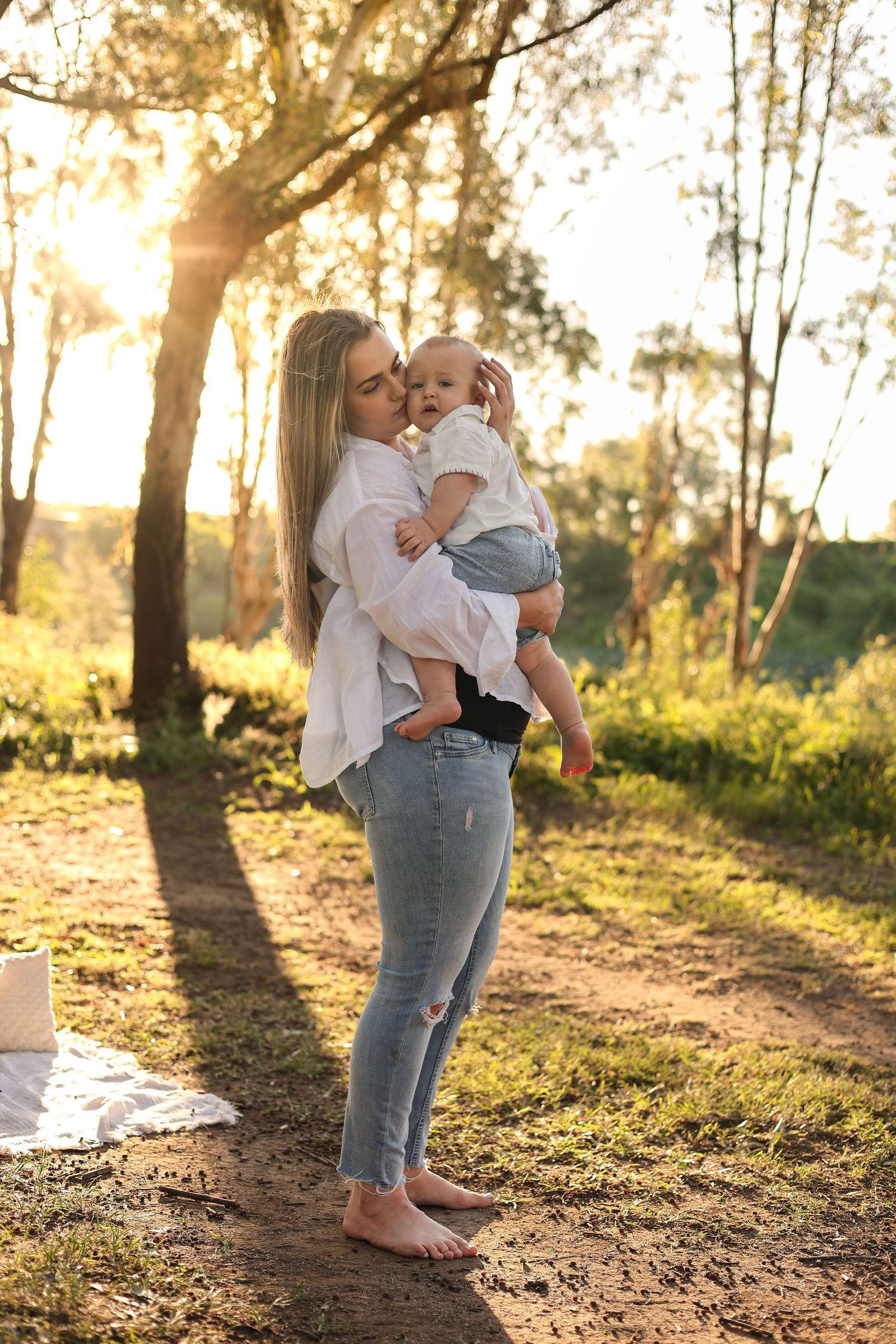 A young mother holds her baby outdoors at golden hour, barefoot in jeans and white shirt, surrounded by sunlit trees.