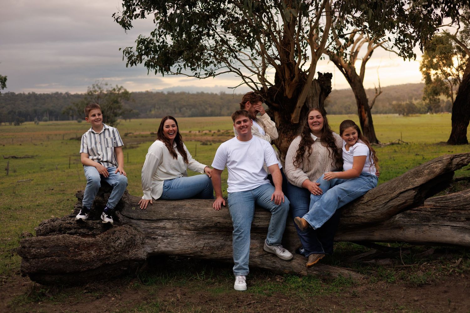 Six people pose on a fallen log in a lush green Australian countryside at dusk, wearing casual jeans outfits.