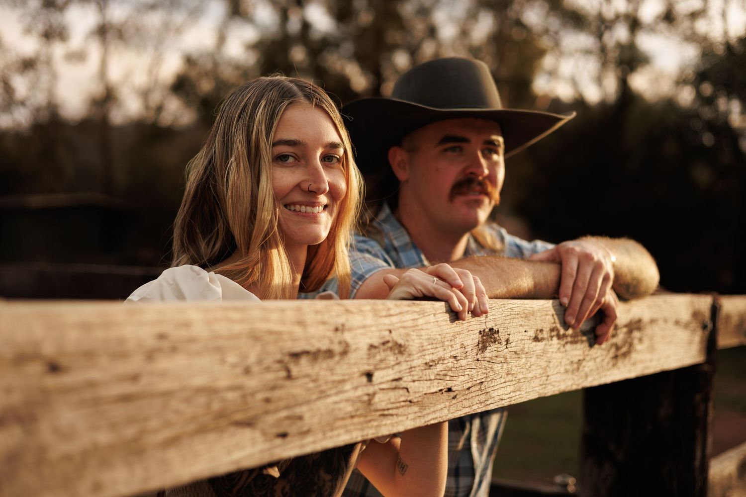 A smiling young woman and a man in a cowboy hat lean on a rustic wooden fence at golden hour on a farm.