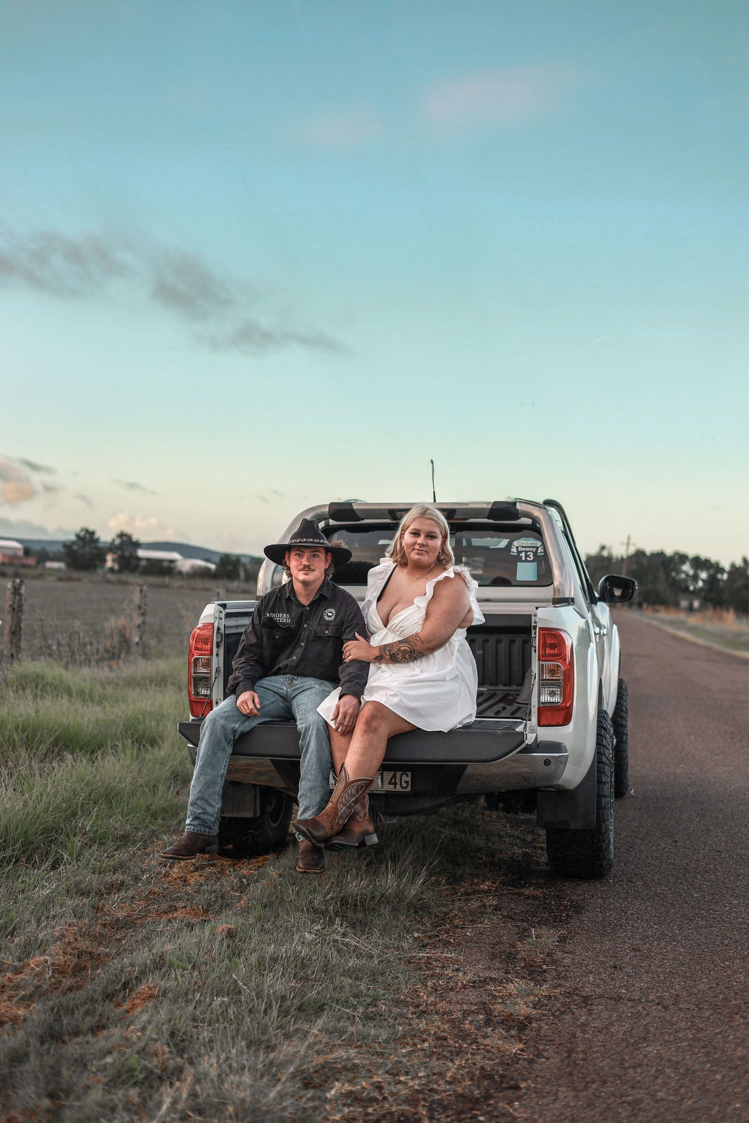 A couple sits together on the tailgate of a pickup truck parked on a rural road at sunset.