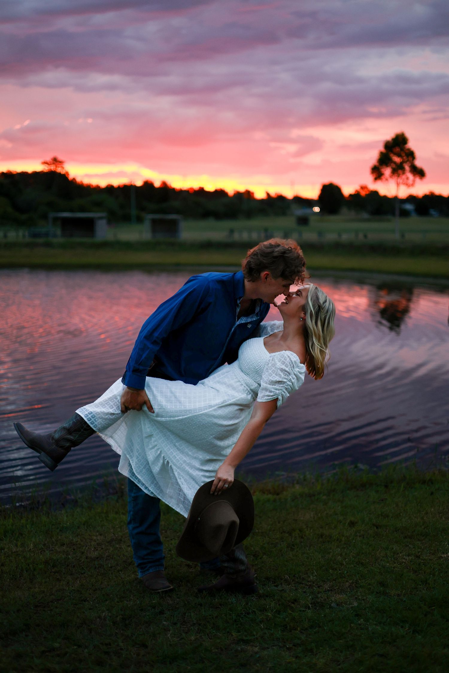 A couple shares a romantic dip pose at sunset by a tranquil lake with vibrant pink and orange sky.