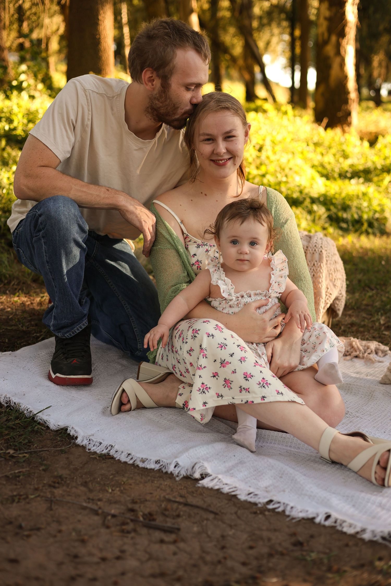 Happy family sitting on blanket in park, man kissing woman's cheek while holding baby girl in floral dress.