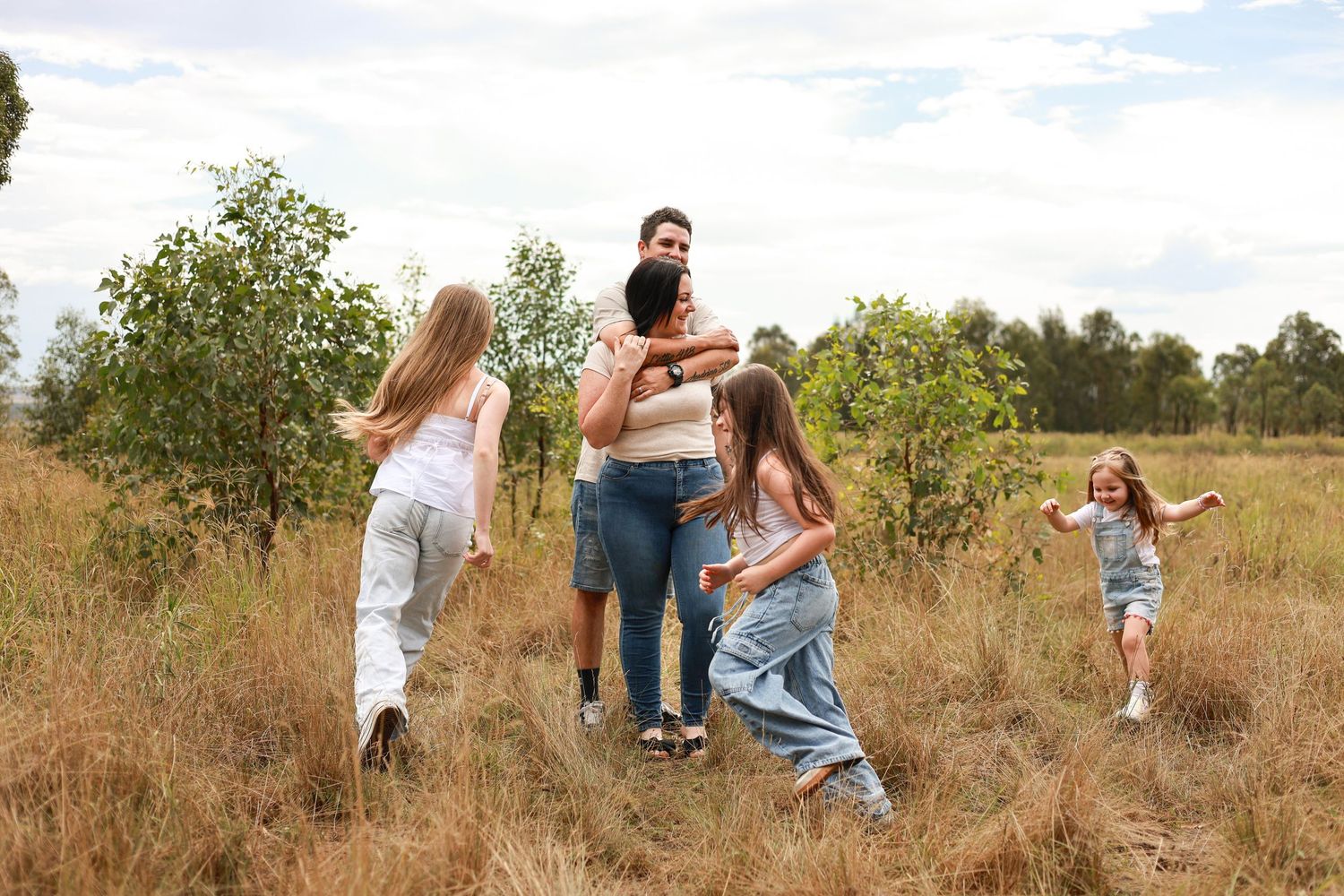 Family with three girls playing in a grassy field surrounded by trees on a cloudy day.