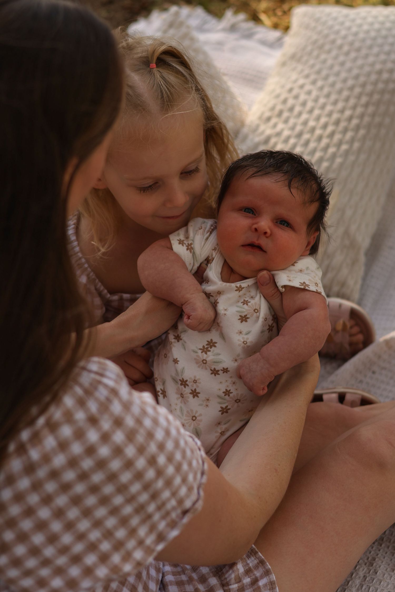 A young girl tenderly holds a newborn baby in a floral onesie, assisted by a woman on a cozy blanket.