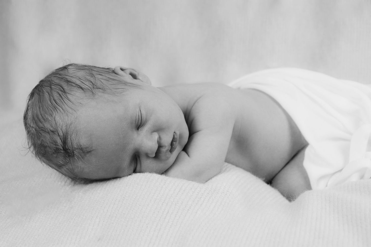 A peaceful sleeping newborn baby lying on soft white bedding in black and white.