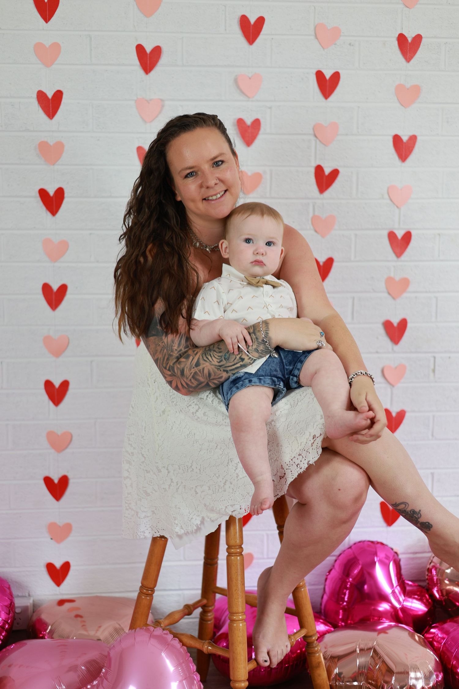 Sweet Valentine's photo with pink and red heart backdrop, pink balloons, and a white dress against painted brick wall.