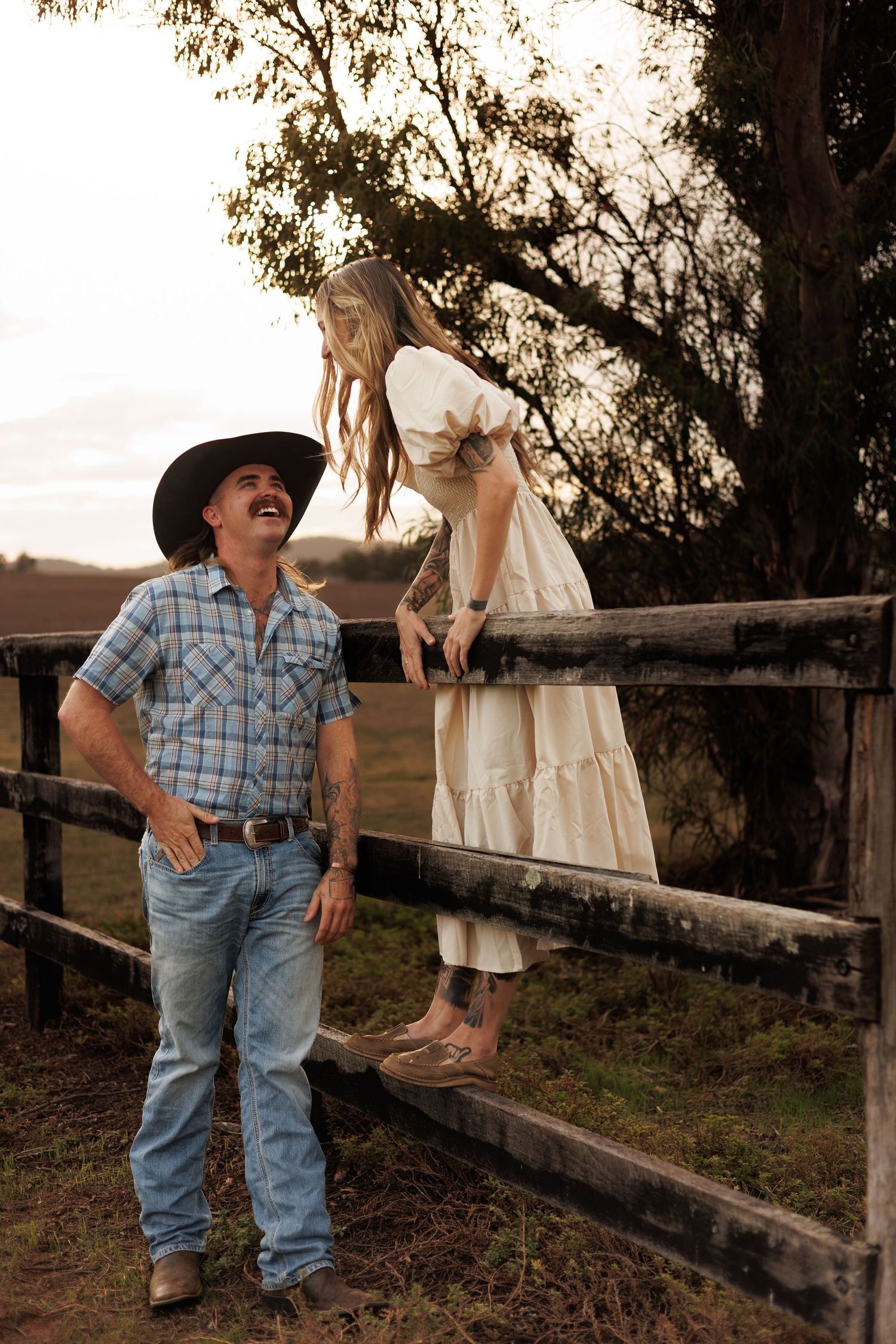 A cowboy in a hat and woman in a white dress laugh together by a rustic wooden fence at golden hour.