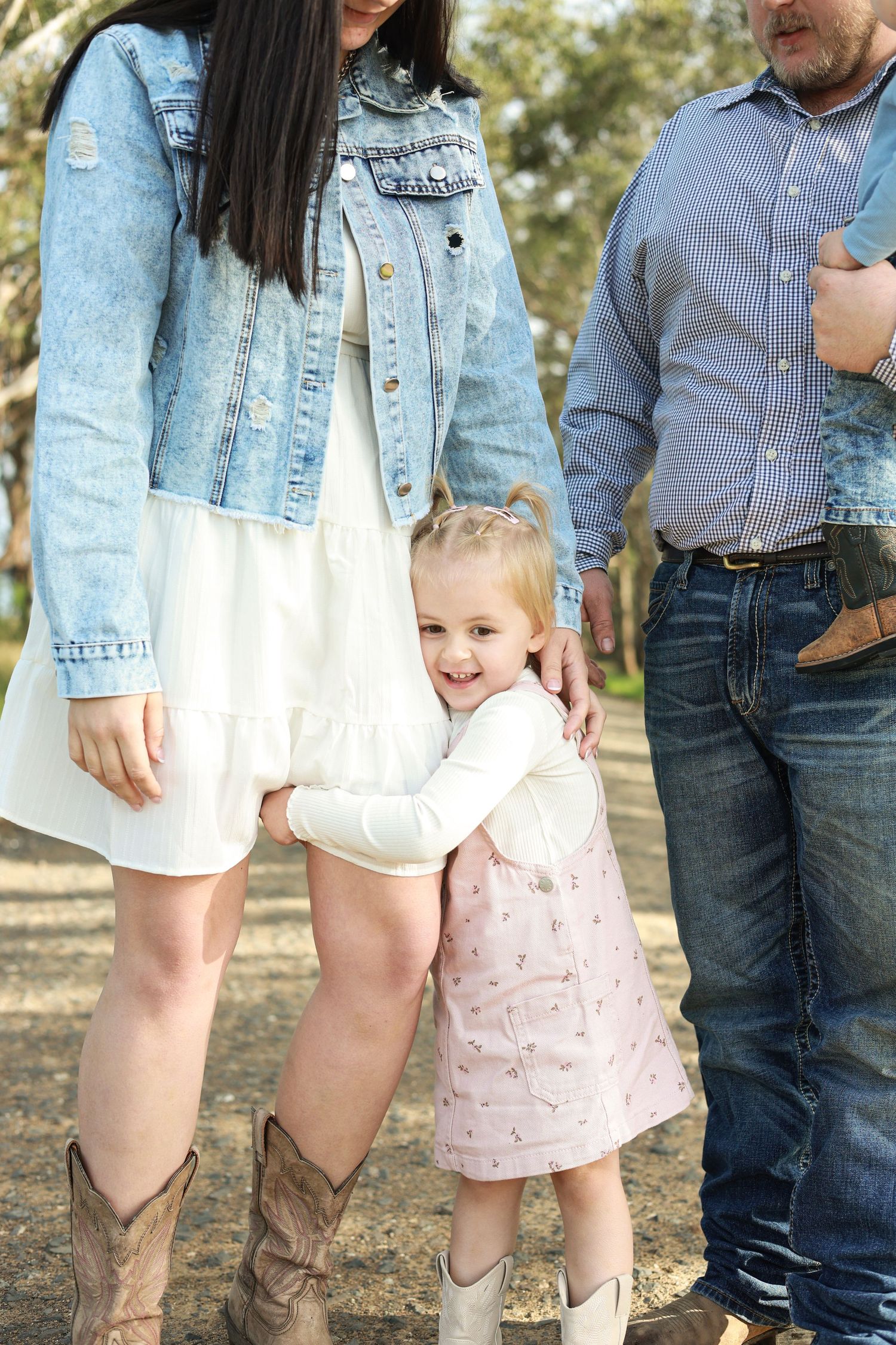 A young girl in a pink dress hugs her mother's legs outdoors, as parents in denim stand nearby.