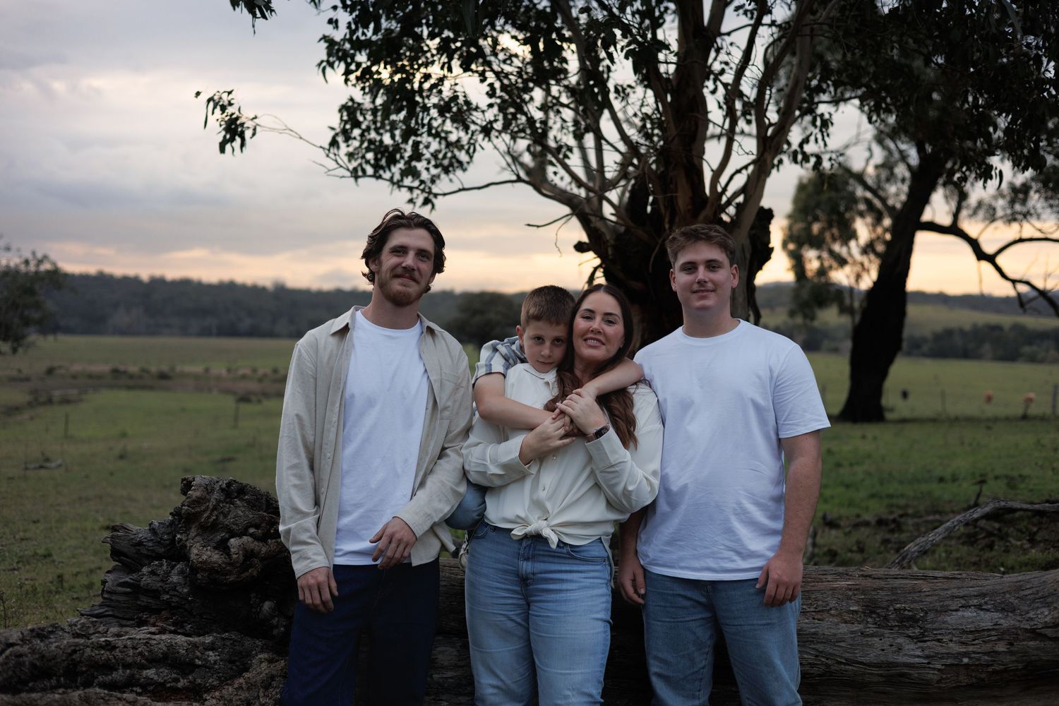 Family of four posing outdoors at sunset near a tree with green fields in the background.