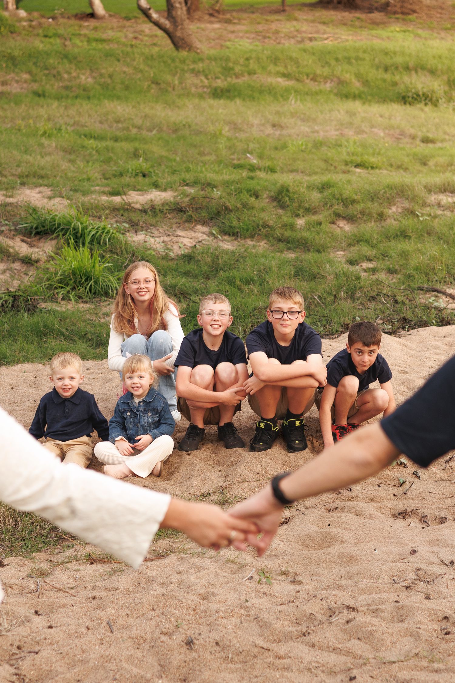 Family group photo with children sitting on sandy ground, smiling at camera in a grassy outdoor setting.