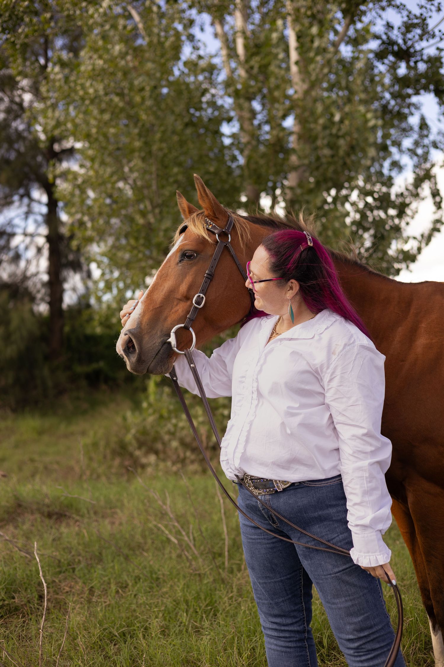 A person in a white shirt and jeans stands beside a chestnut brown horse in a grassy field.