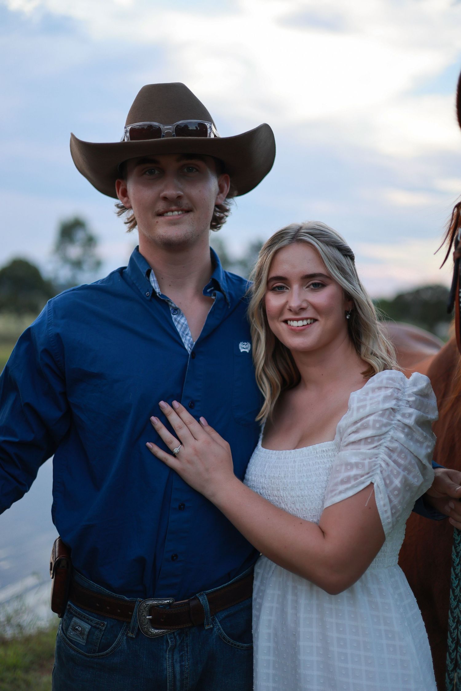 A couple poses outdoors at sunset, with the person in western attire and cowboy hat next to someone in a white dress.