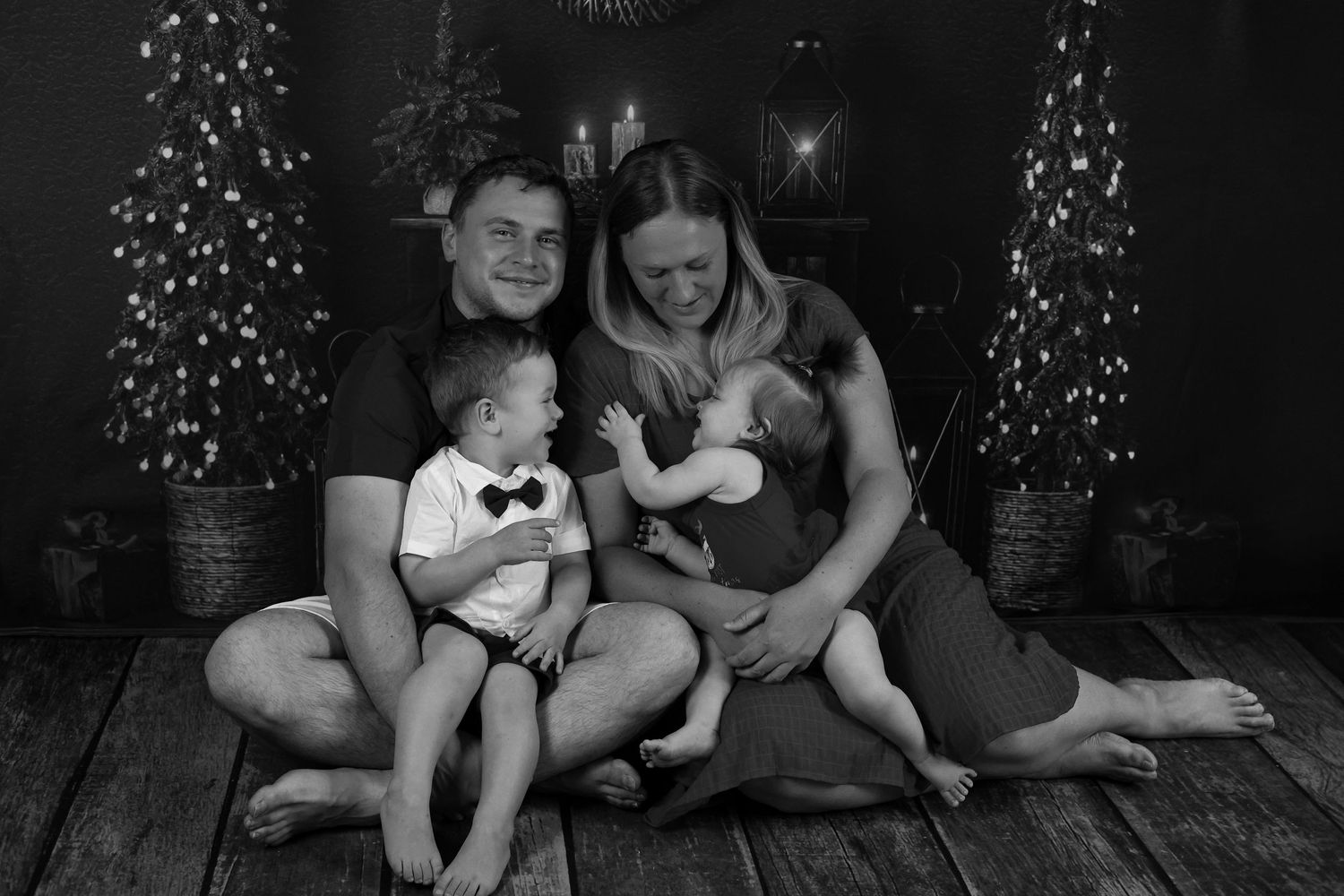 A heartwarming black and white holiday portrait of a family sitting together between decorated Christmas trees.