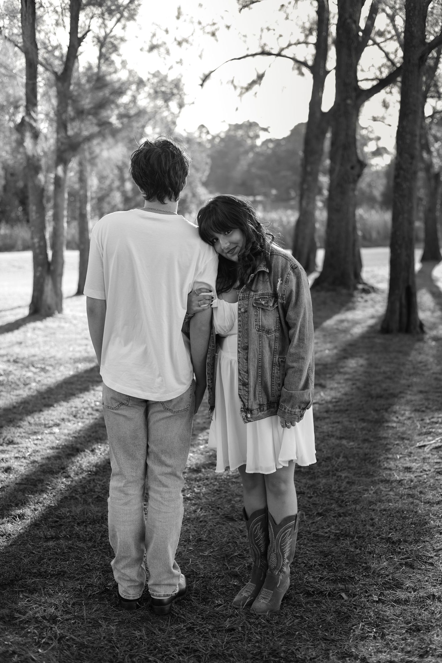 Black and white photo of a couple standing in a park, woman in denim jacket and cowboy boots leaning on mans shoulder.