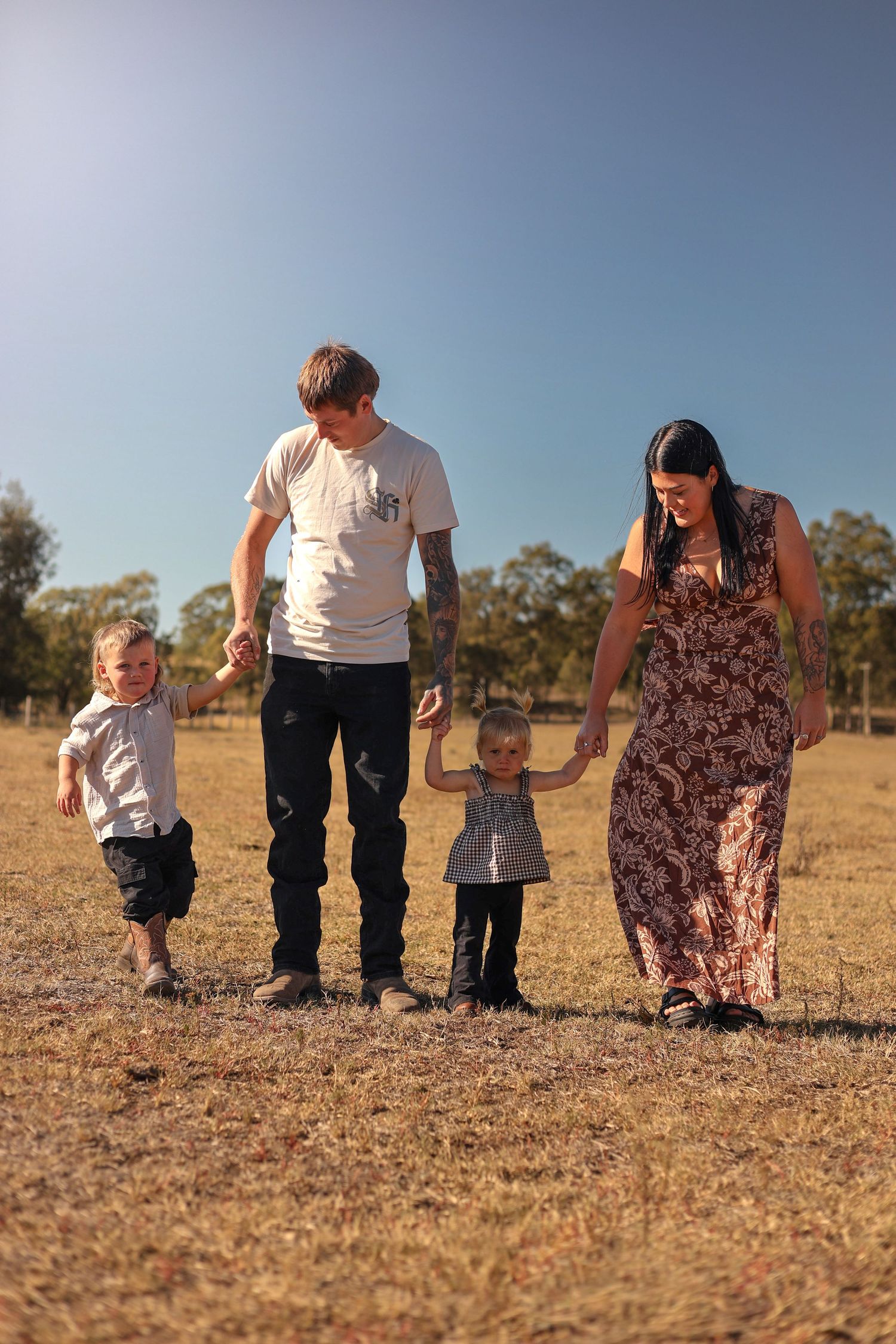Family of four walking hand-in-hand in a sunny field, parents smiling at their two young children.