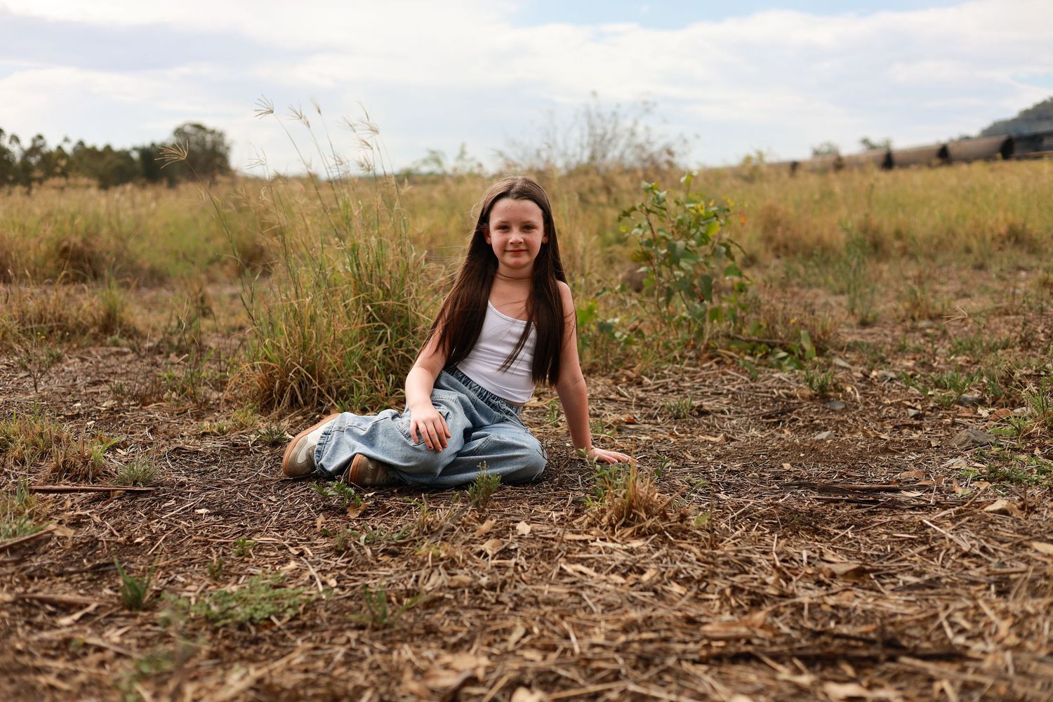 Young girl sitting cross-legged in a dry grassy field, smiling, wearing jeans and a white tank top.