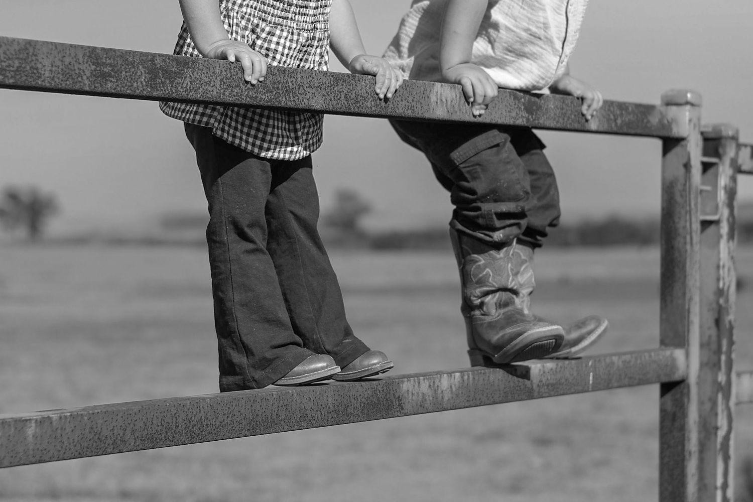 Two children standing on a metal farm gate in cowboy boots, black and white rural countryside photo.