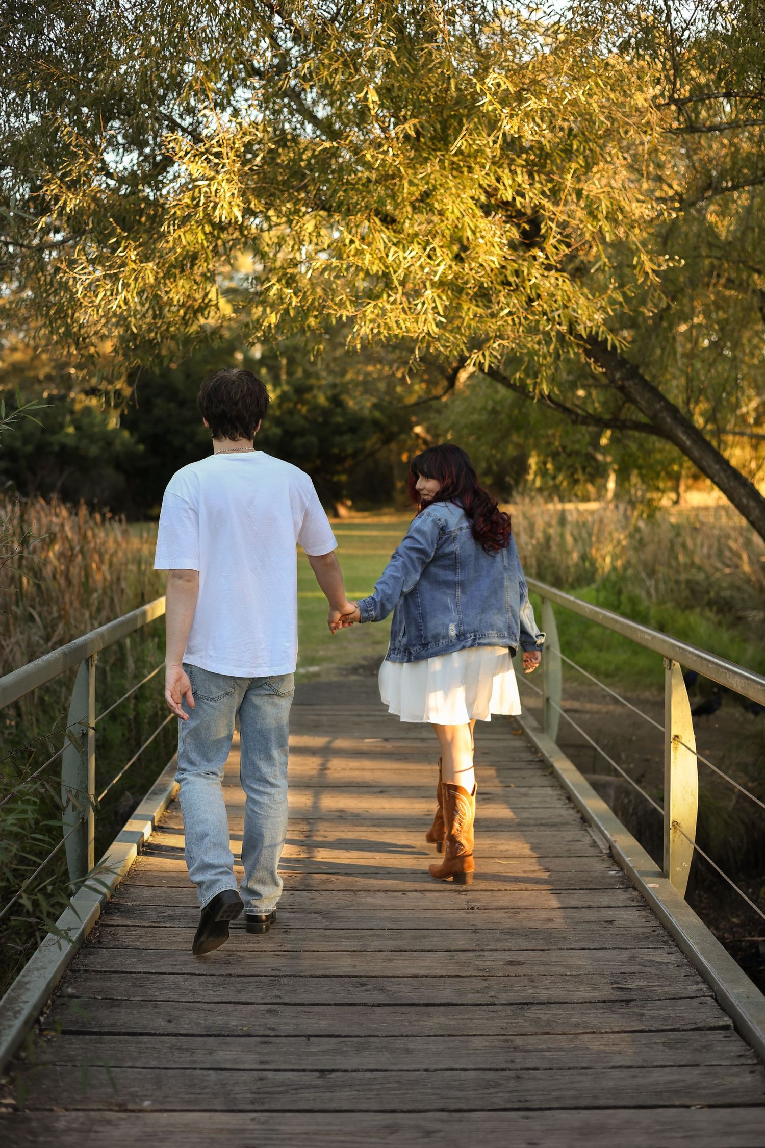 Couple holding hands walking across a wooden bridge at golden hour sunset in a lush green park.