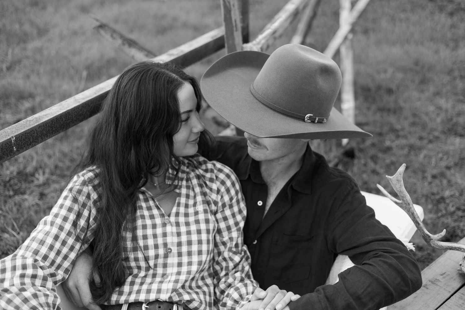 Black and white photo of a couple in western attire sharing a romantic moment outdoors near a wooden fence.