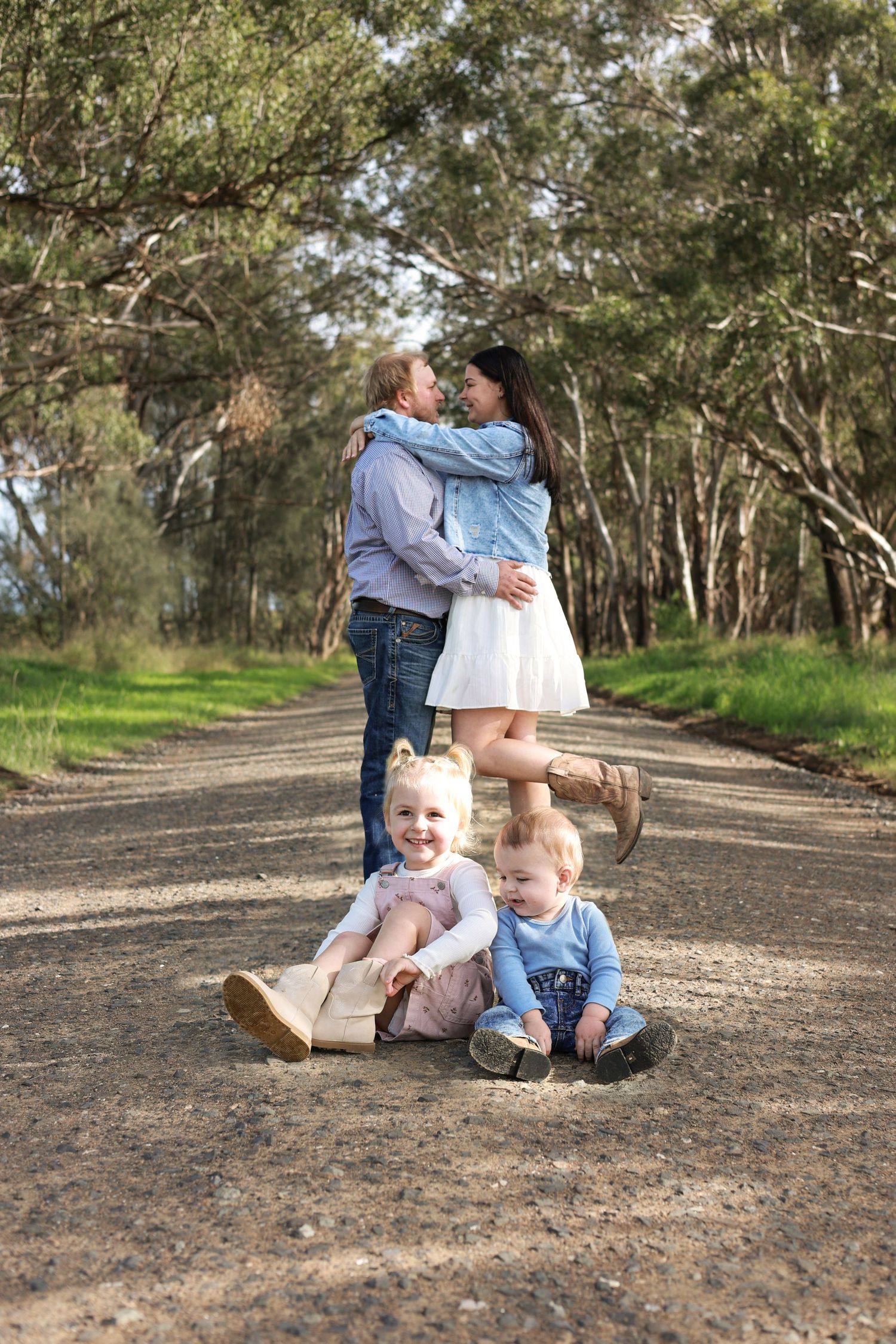 Family photo with parents kissing on a dirt road surrounded by trees while two young children sit smiling in the foreground.