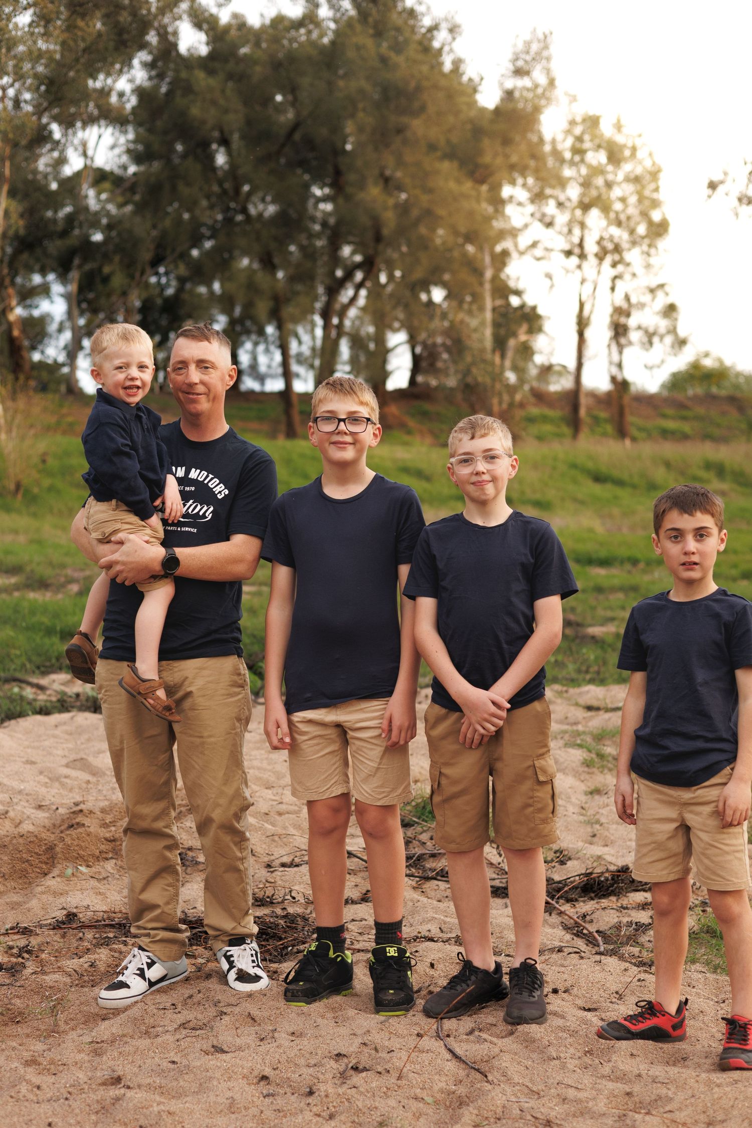 A man holding a toddler poses outdoors with four boys in matching navy shirts and khaki shorts on sandy ground.