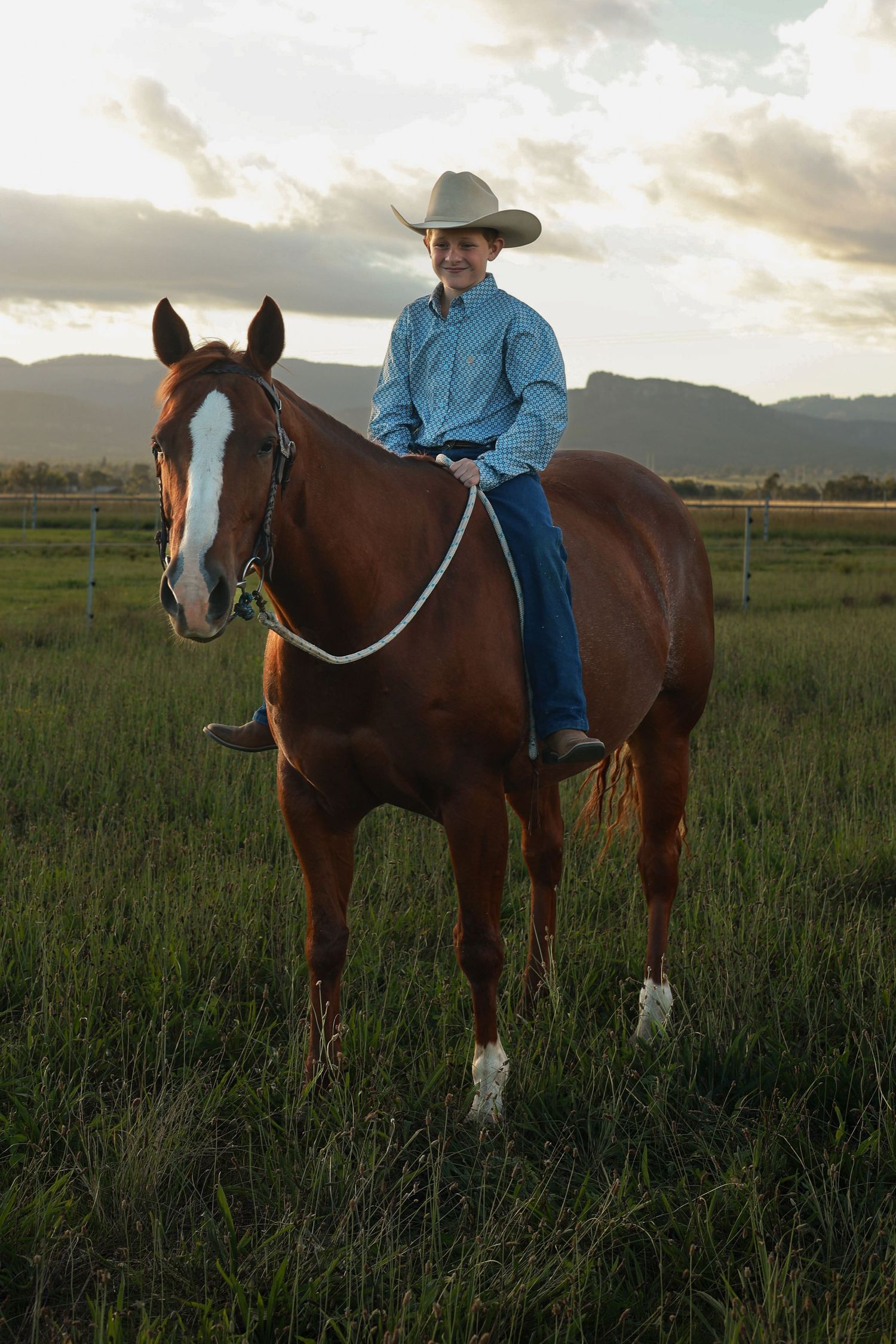 A young cowboy in a blue shirt and white hat rides a chestnut horse in a green field with mountains at sunset.