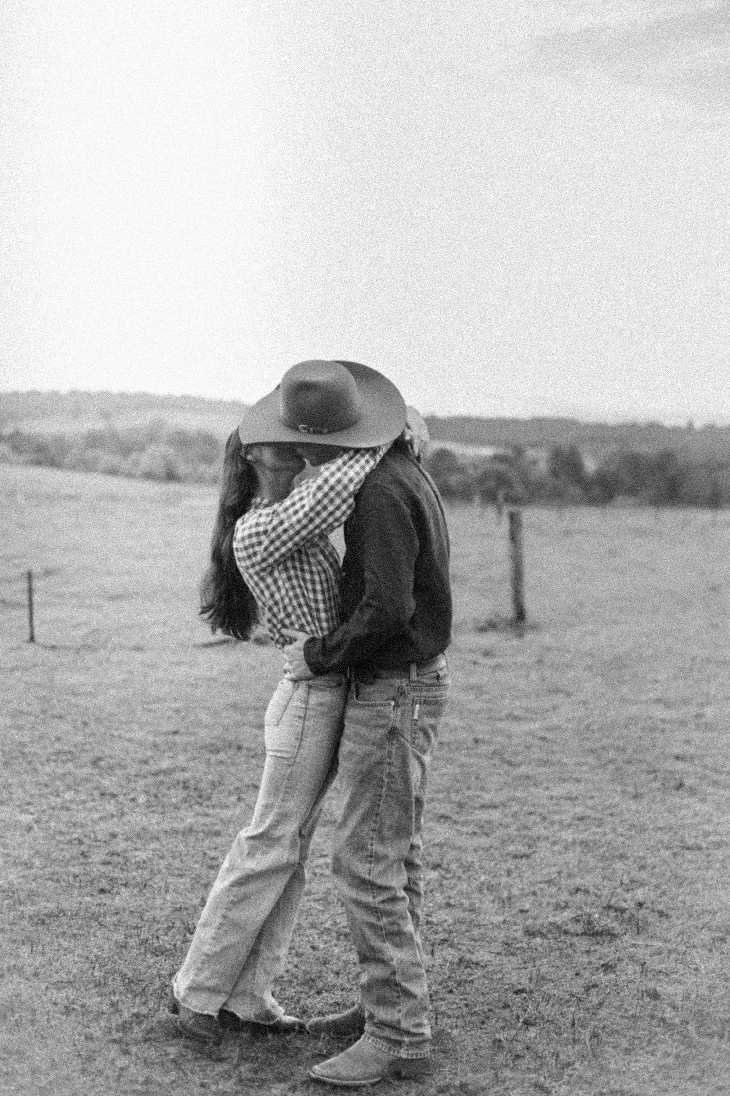 Black and white photo of a couple sharing a kiss in a field, wearing cowboy hats and jeans.