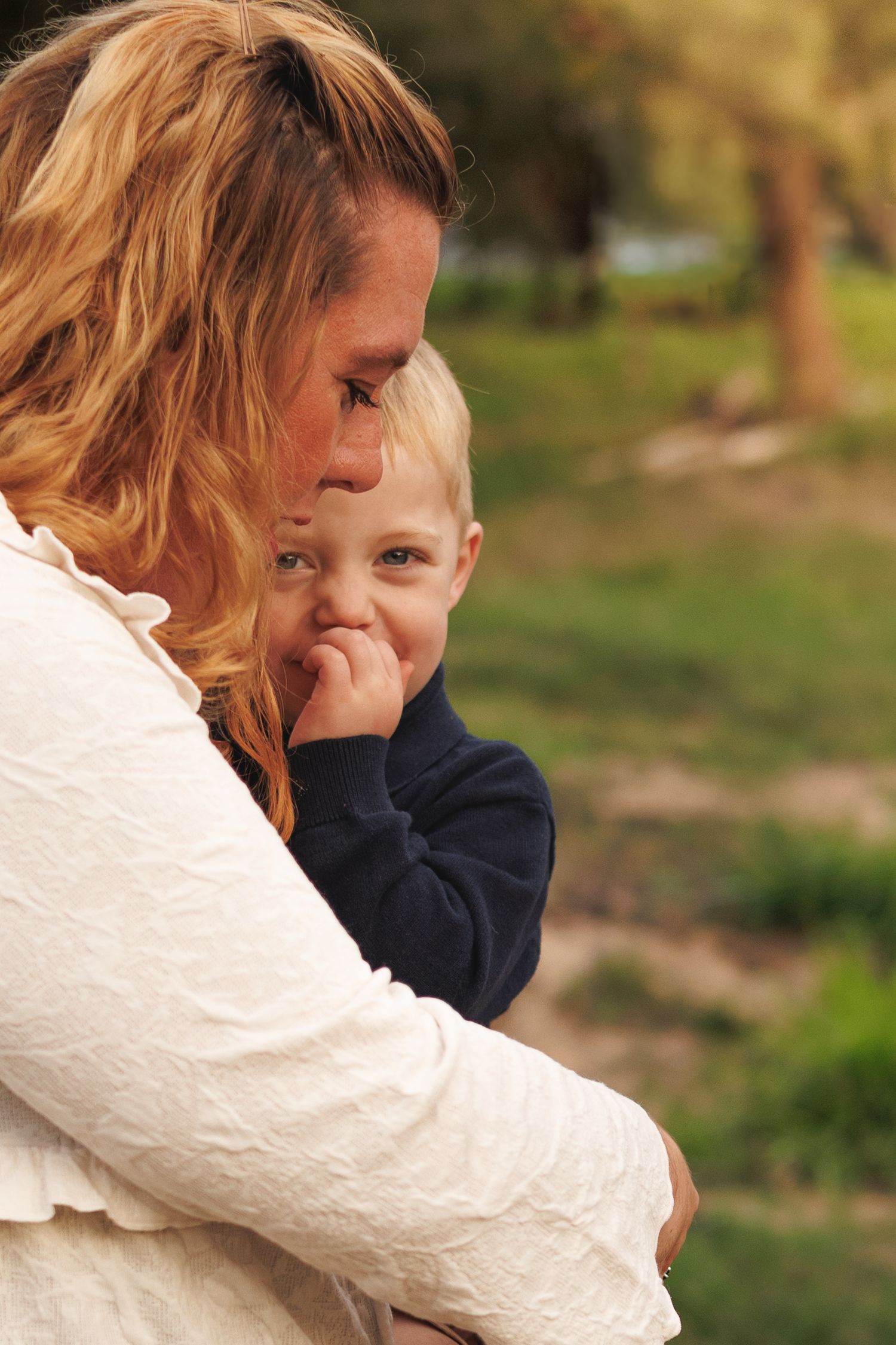 A mother with blonde hair holds her young toddler boy outdoors in a green park setting.