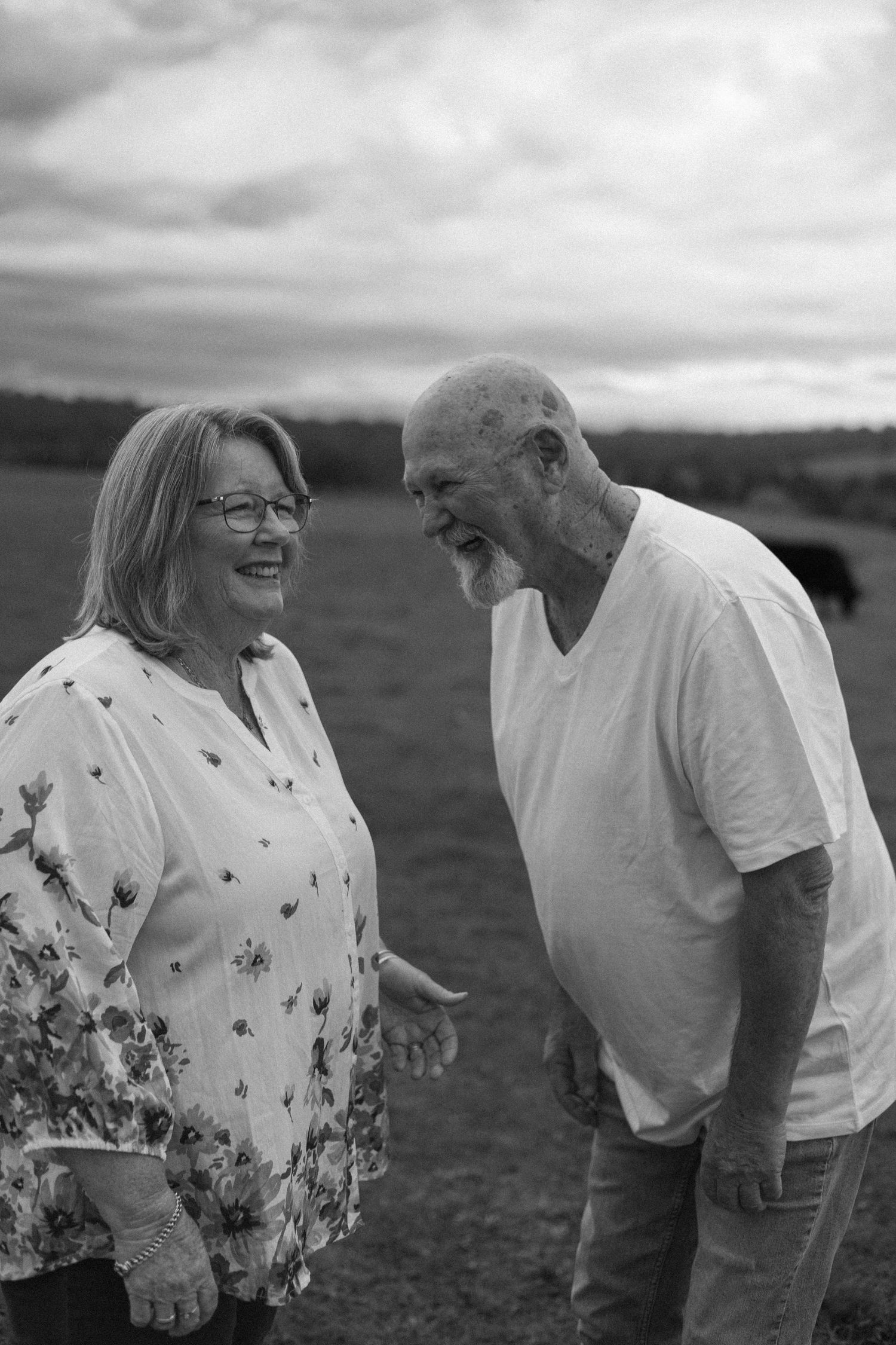 Elderly couple laughing together outdoors in a field, captured in black and white photography.