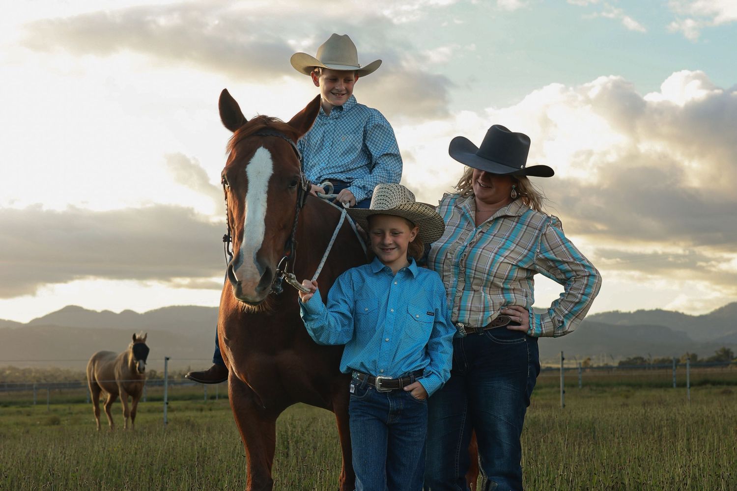 Three cowboys in hats pose with a chestnut horse in a green field at sunset with mountains in the background.