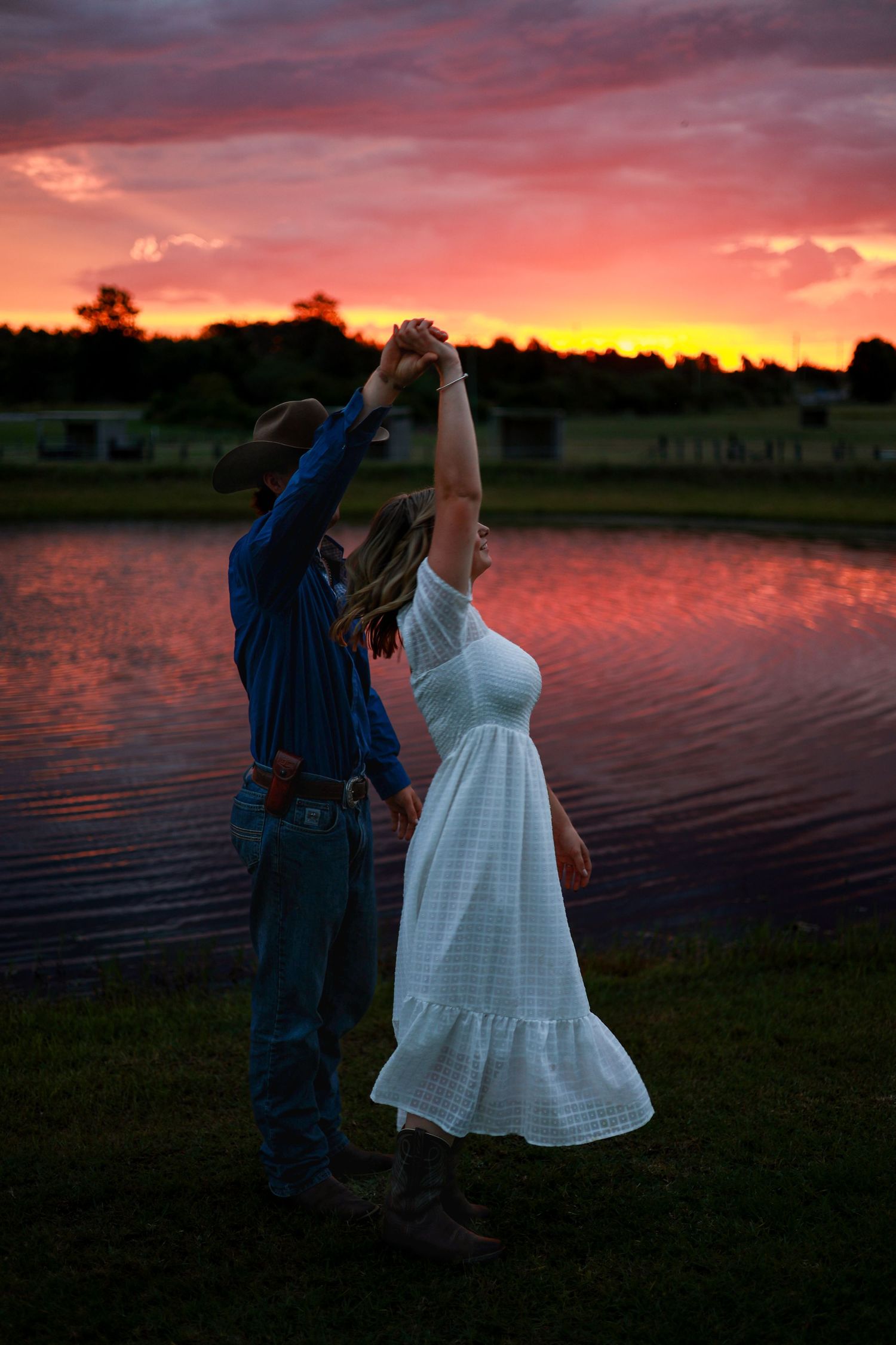 A couple dances by a lake at sunset with vibrant orange and pink skies reflecting on the water's surface.