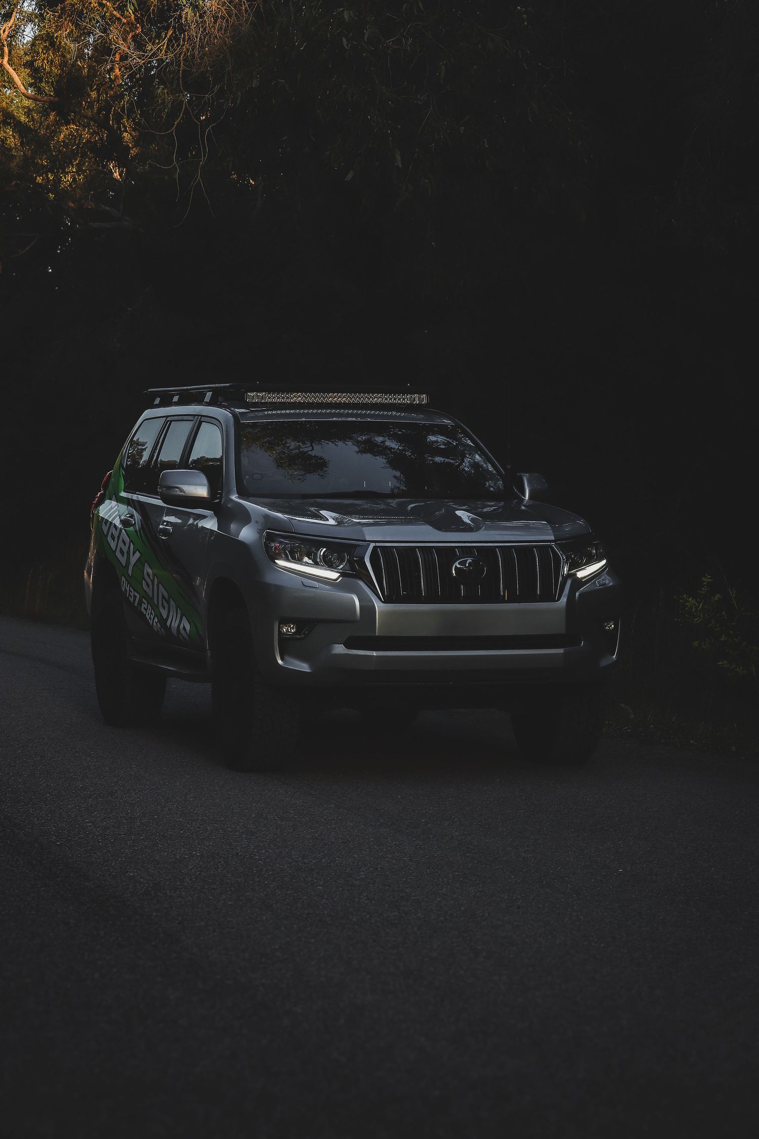 A sleek Toyota Land Cruiser Prado SUV parked in shadows with its LED headlights illuminated against a dark background.