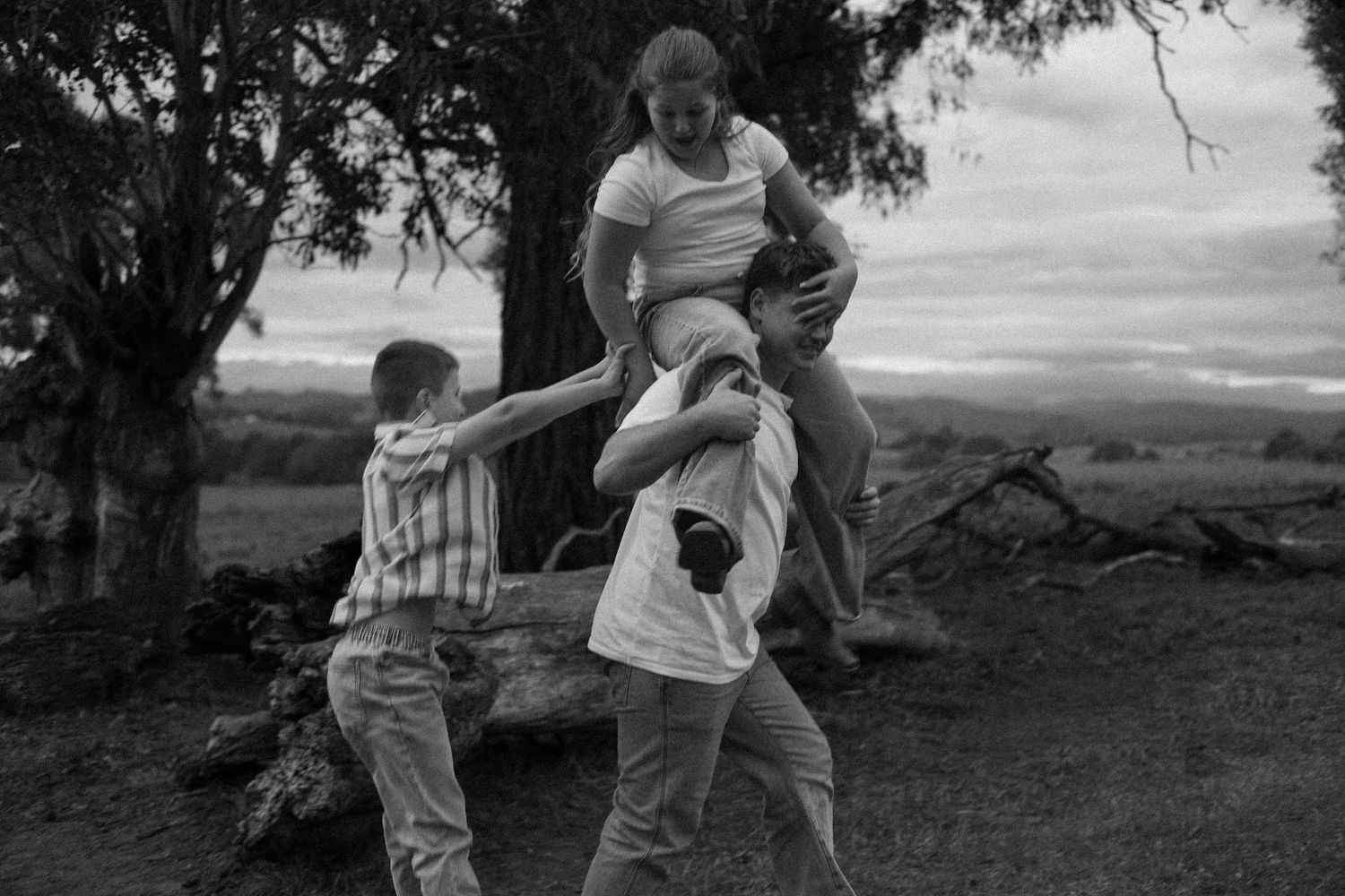 Three children playing outdoors in black and white, one being lifted up near a large tree in a rural landscape.