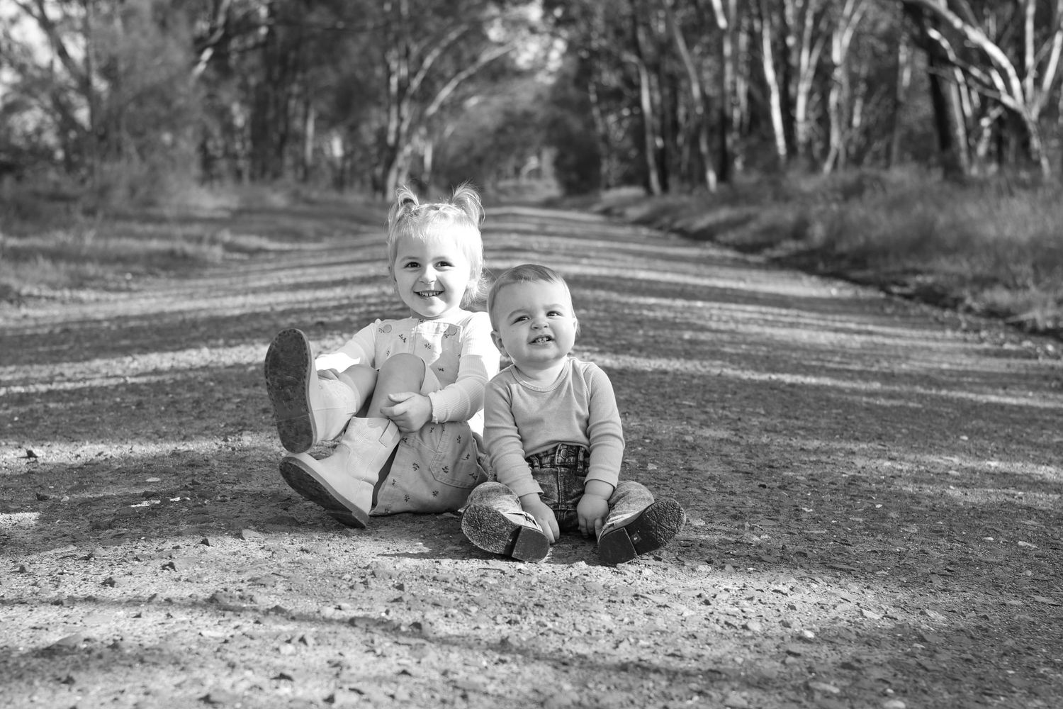 Two happy toddlers sitting together on a dirt road surrounded by trees in a black and white photo.