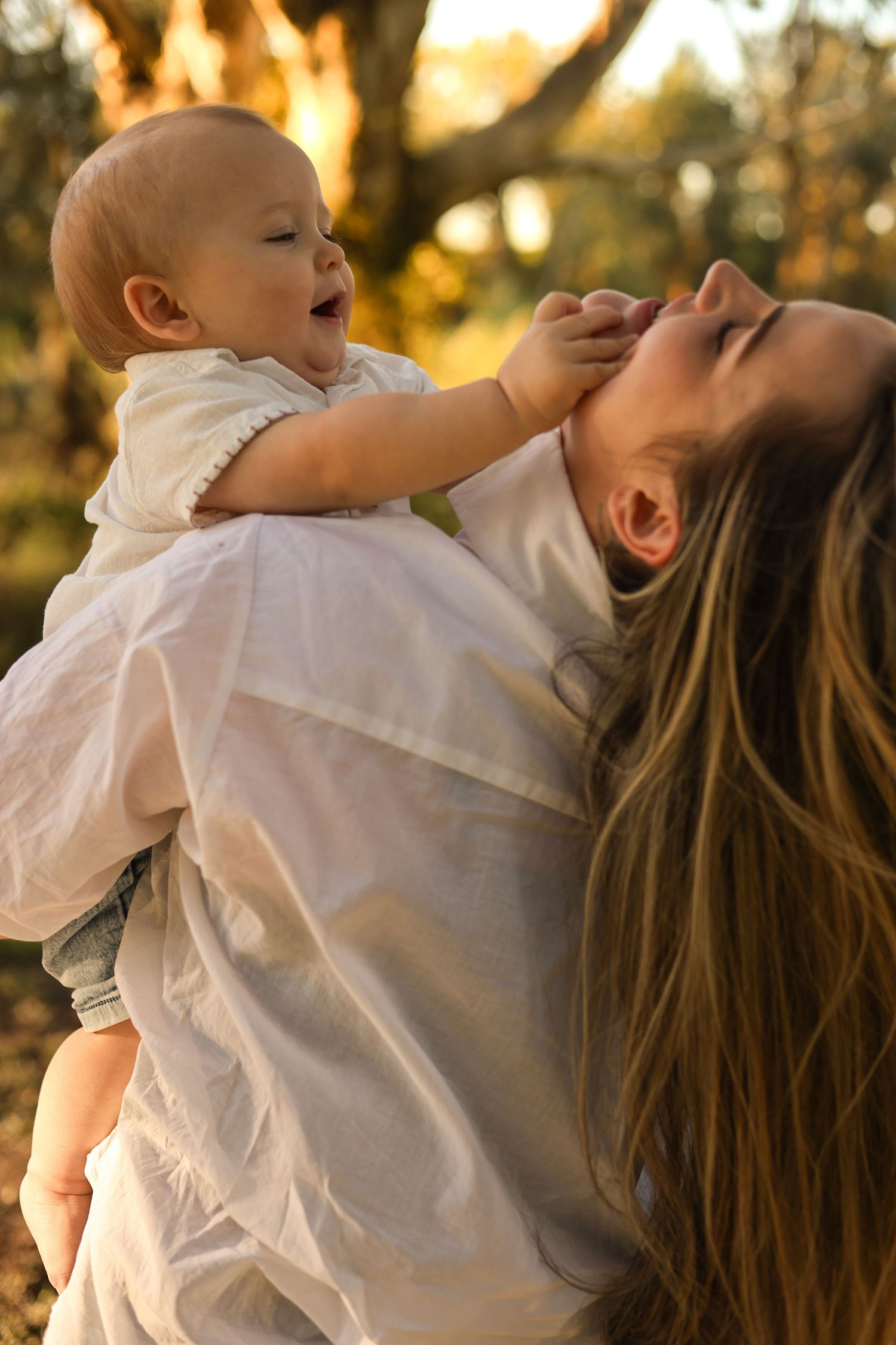 A joyful mother holds her laughing baby outdoors in golden sunset light, both wearing white outfits.