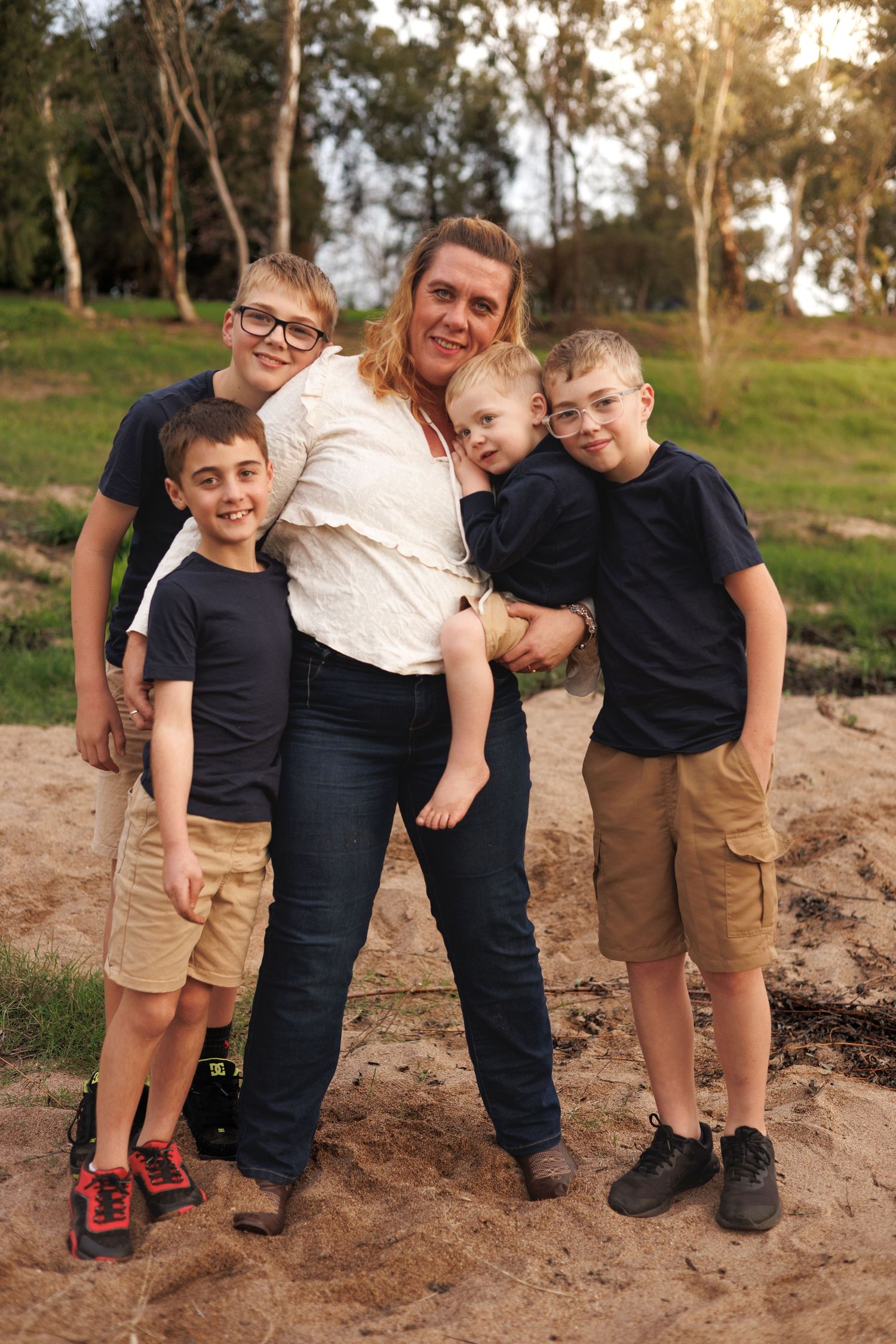 A mother poses outdoors with her four young sons on sandy ground with green trees in the background.