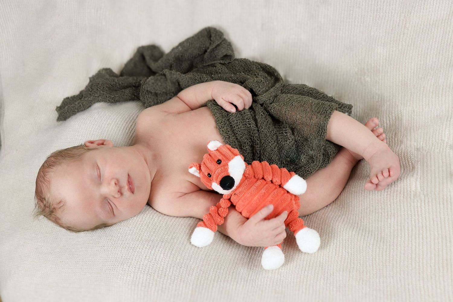 Sleeping baby cuddling with orange stuffed toy fox on light colored blanket.